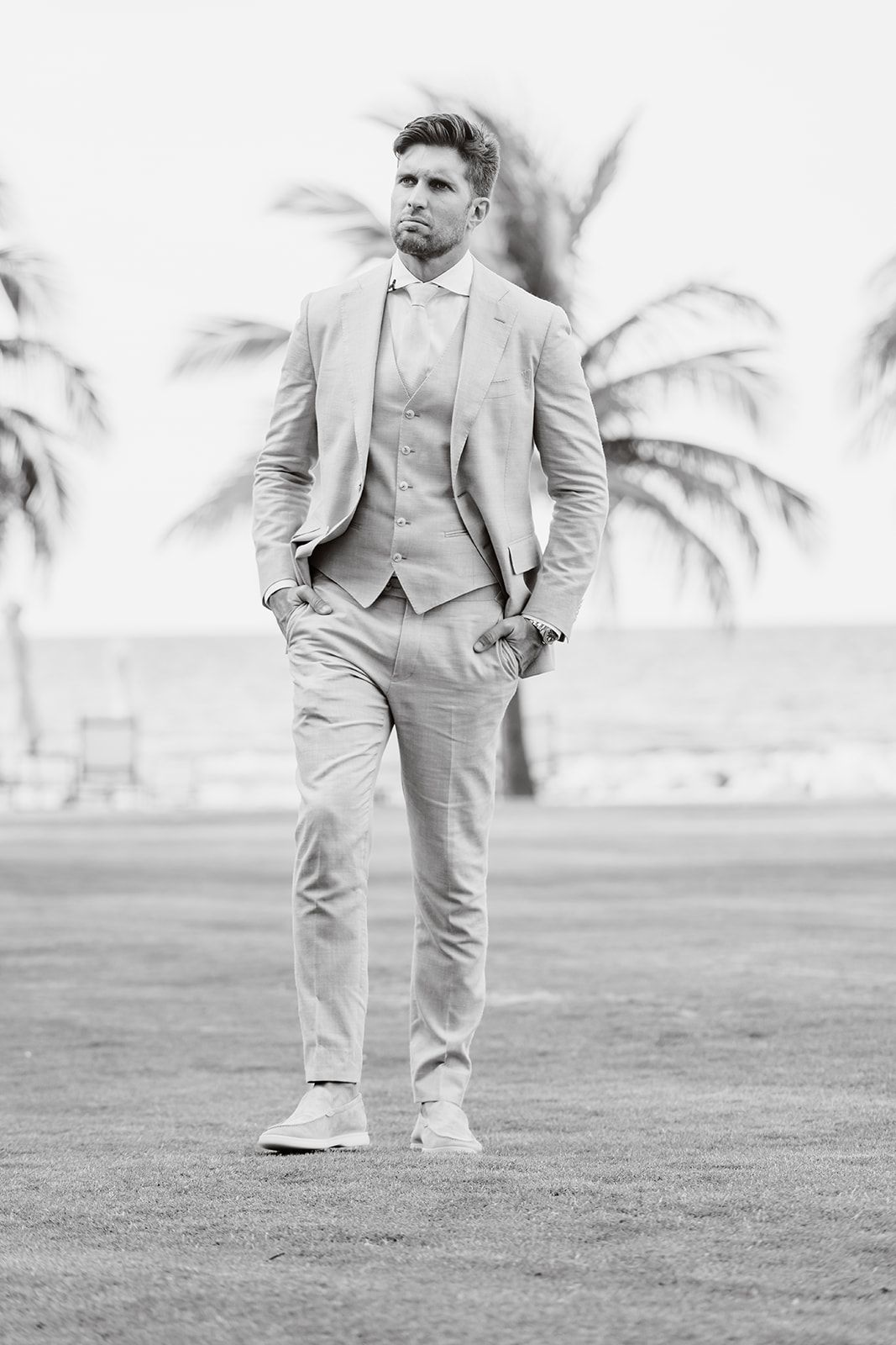 A groom in a light gray three-piece suit stands confidently against a tropical palm backdrop.