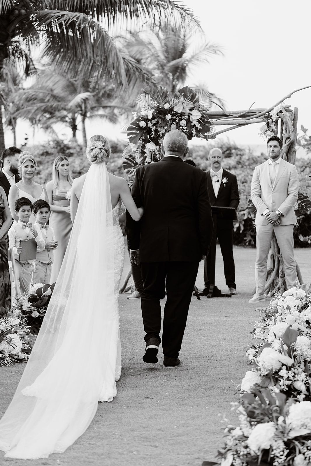 A wedding ceremony under palm trees with guests watching the bride walk down a flower-lined aisle.