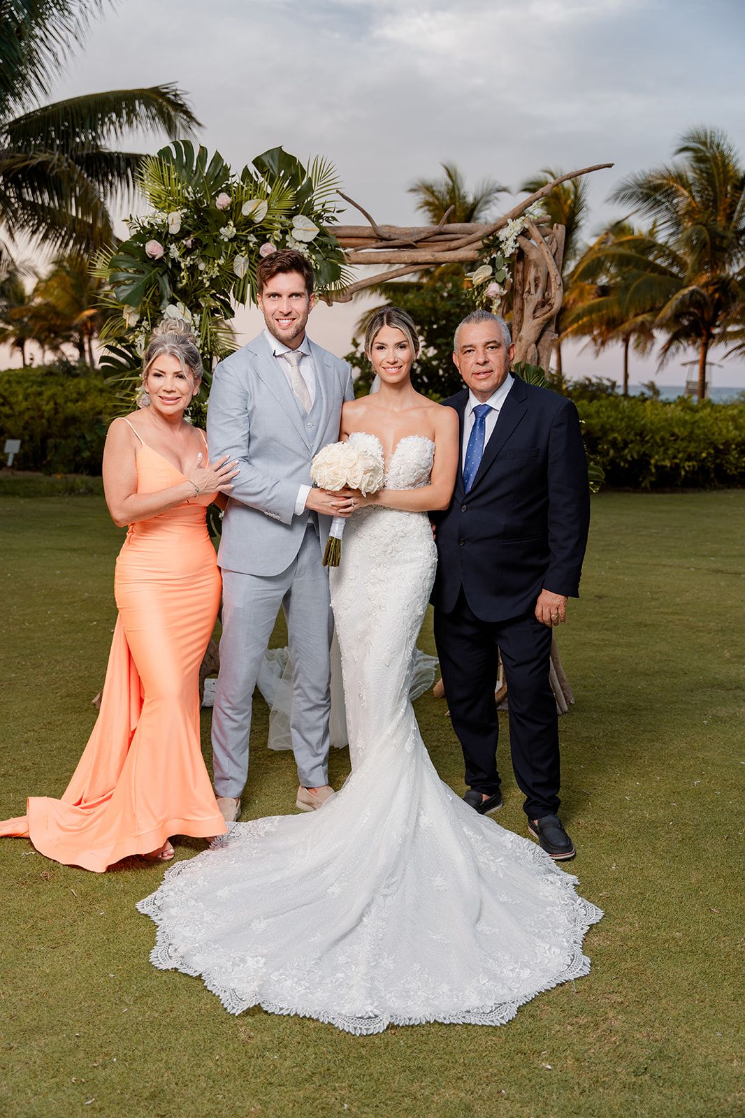 A wedding party poses on a green lawn with palm trees at a tropical beach venue.