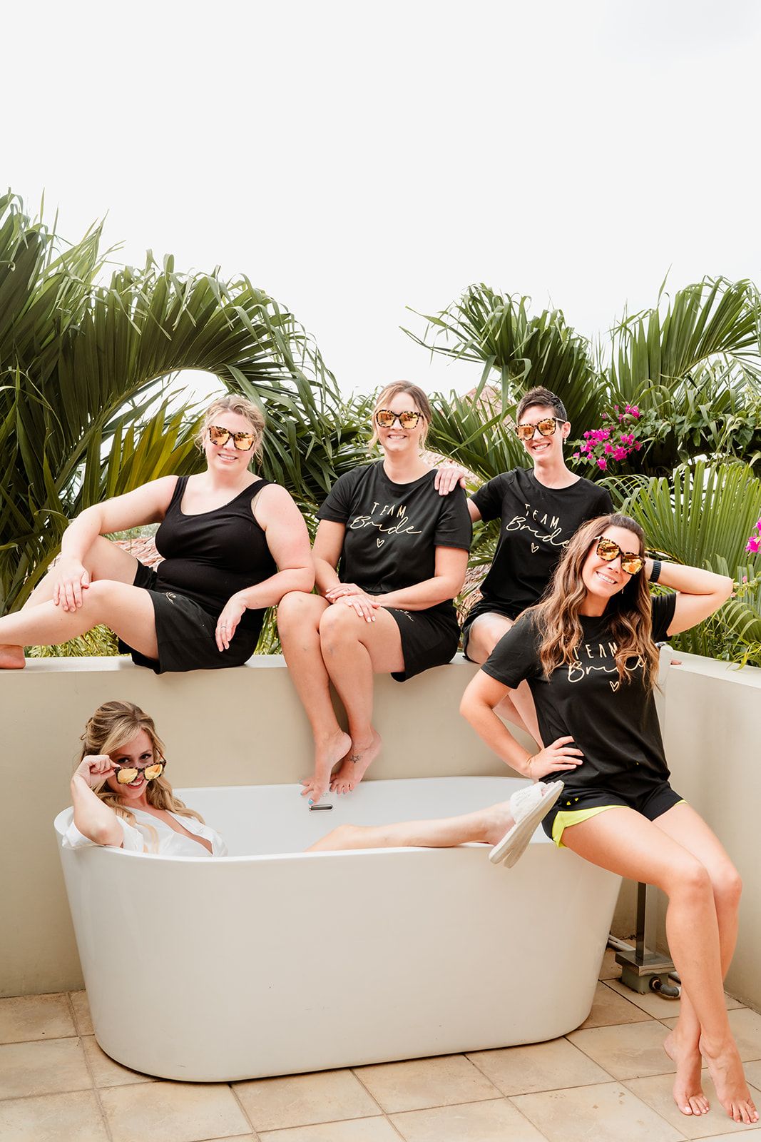 A group in matching black outfits poses together playfully on a white bathtub outdoors with tropical plants in the background.
