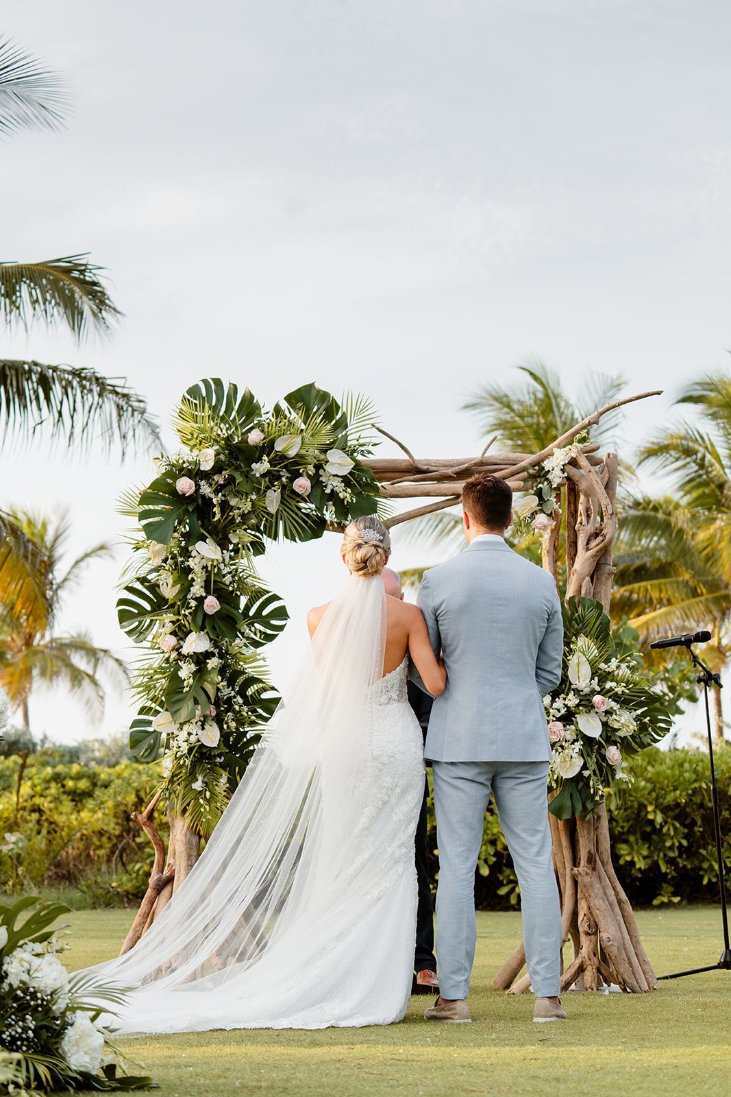 Newlyweds stand beneath a tropical wedding arch adorned with monstera leaves and white flowers.