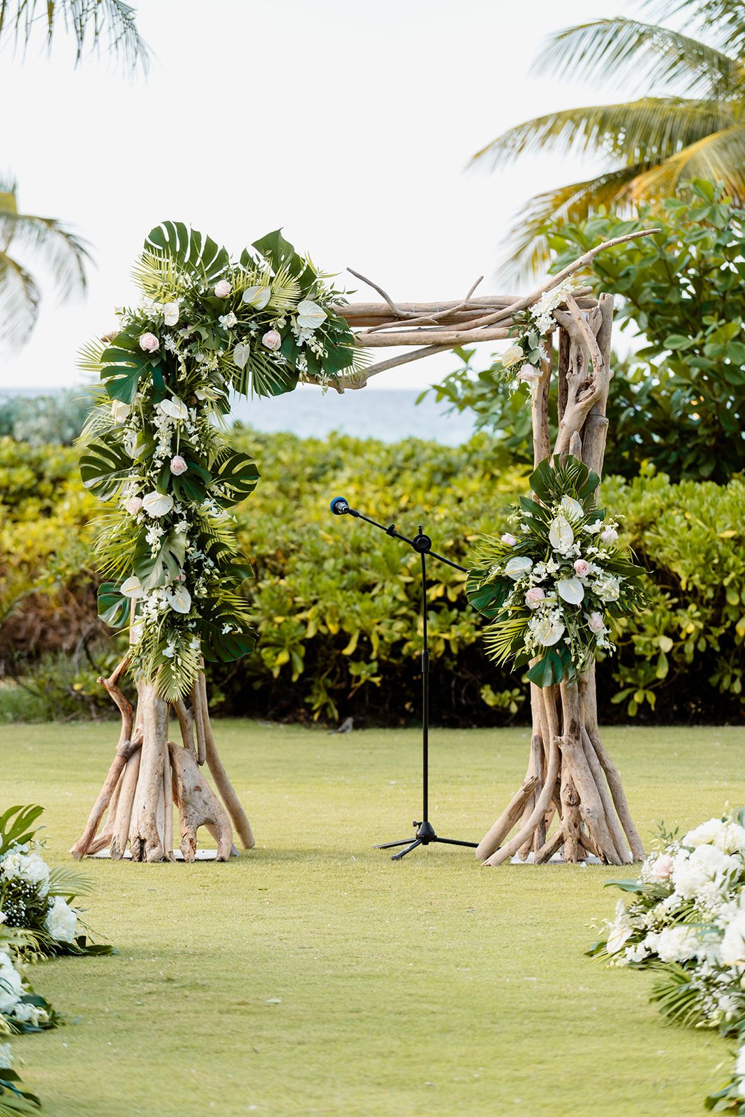Rustic wooden wedding arch decorated with tropical greenery and white flowers on grass lawn.
