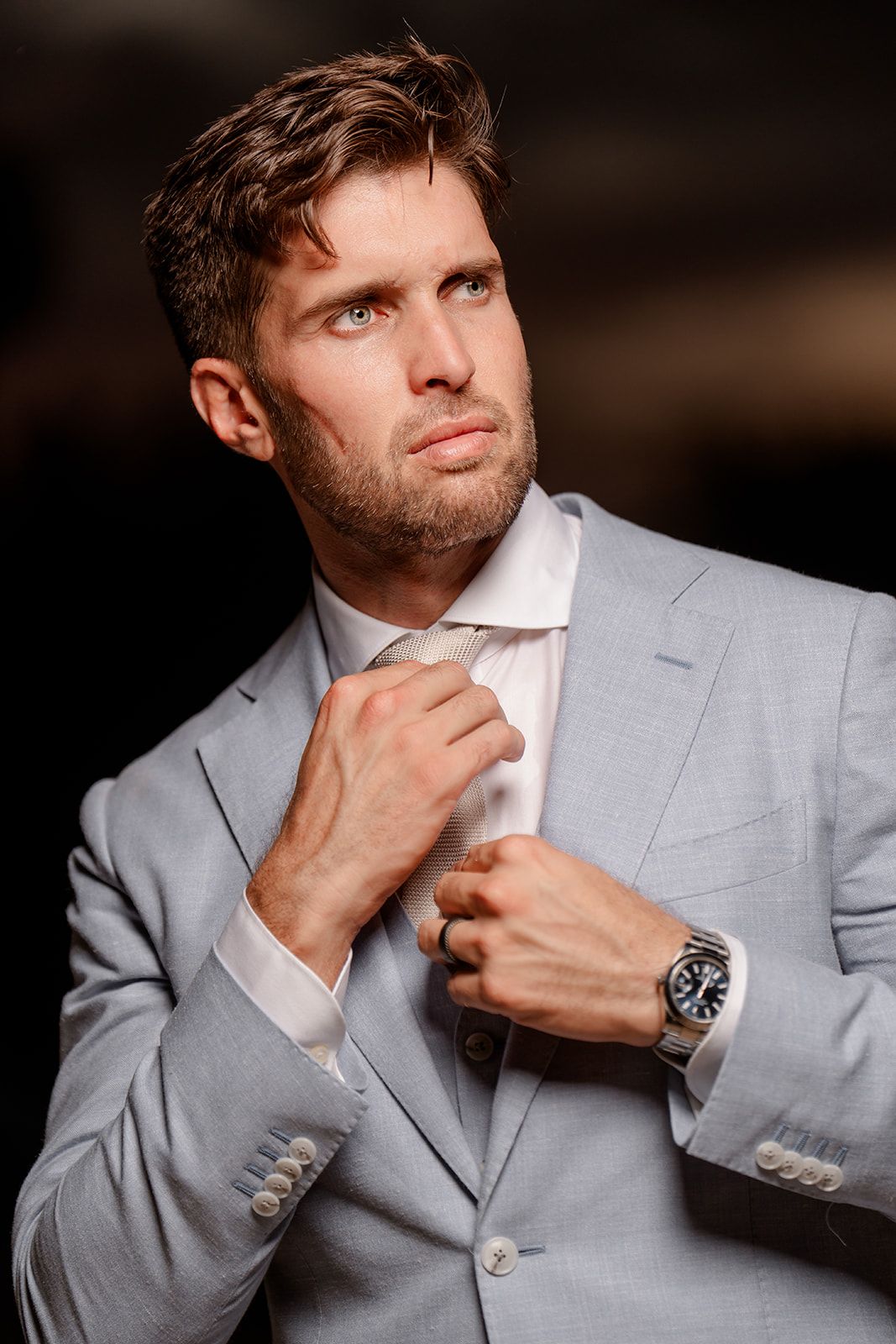Groom in light grey suit adjusts his tie while wearing luxury watch.