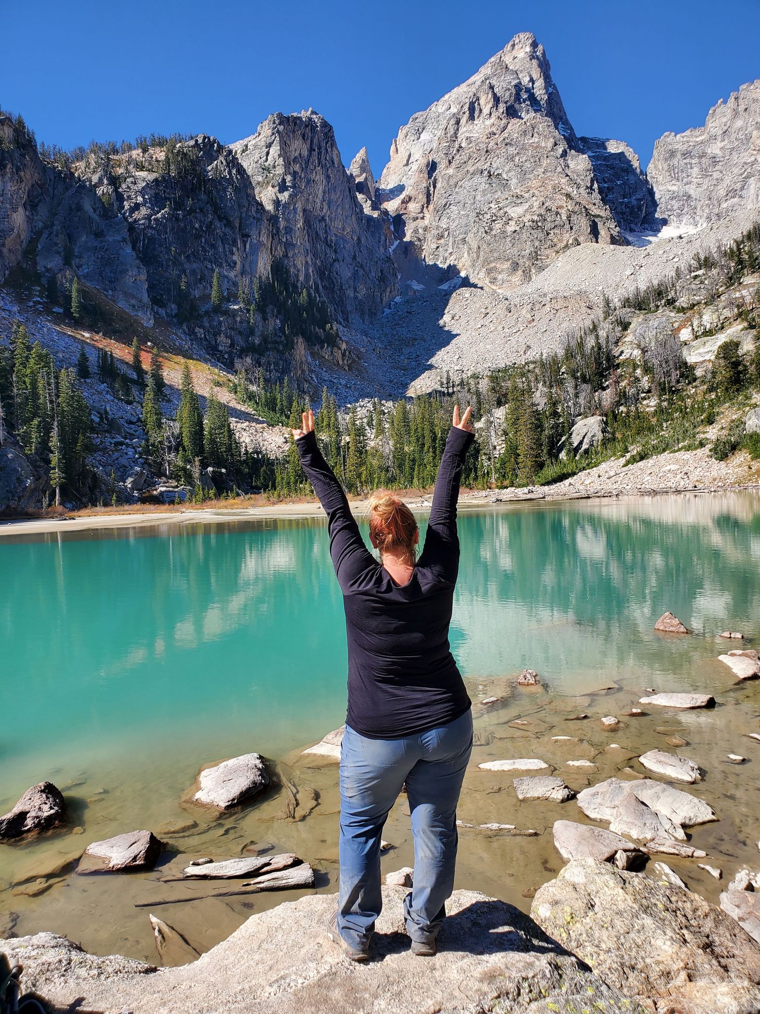 A person celebrates with raised arms at a turquoise alpine lake surrounded by rocky mountain peaks in stunning wilderness.