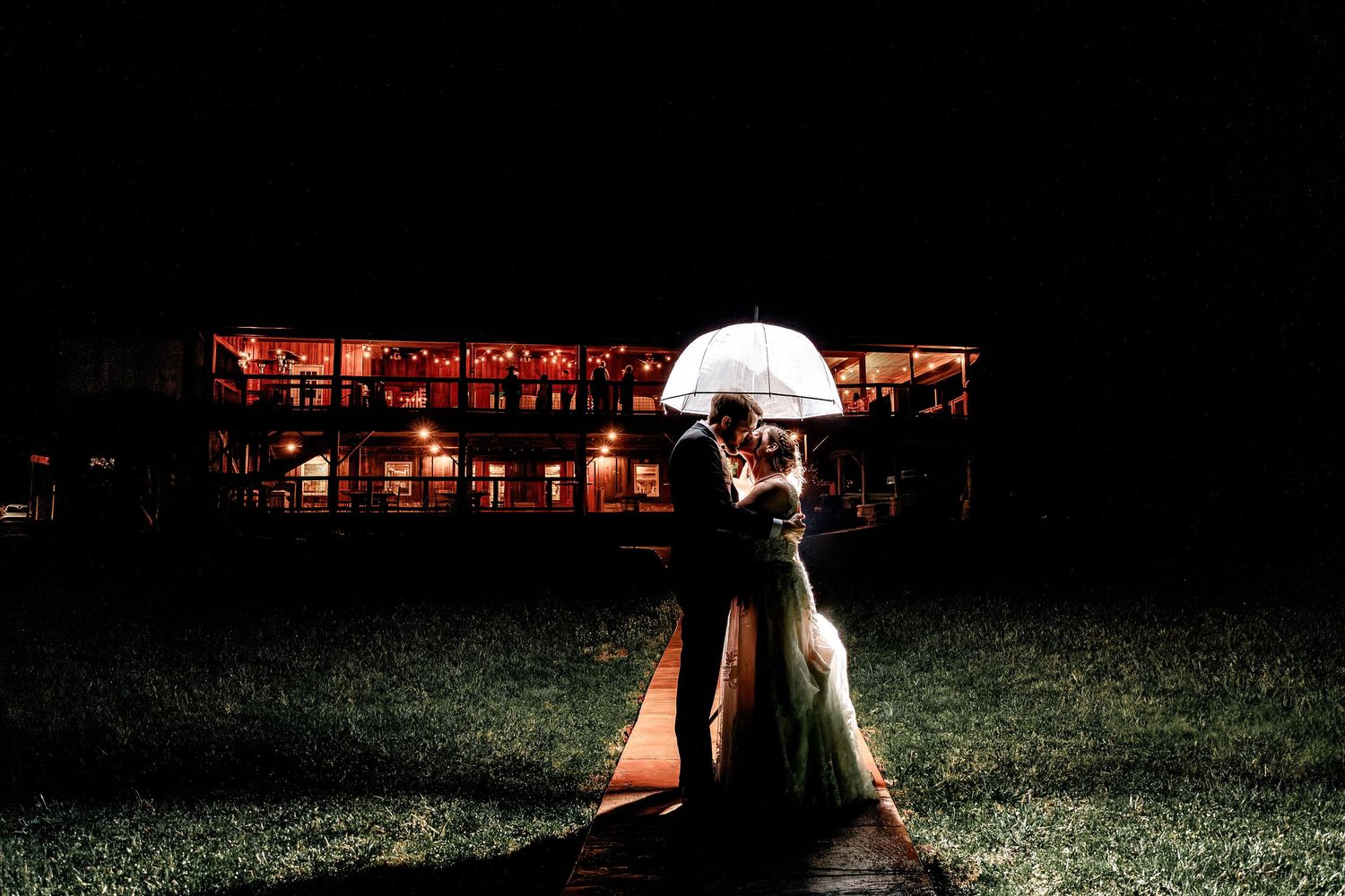 Romantic nighttime wedding photo of bride and groom under umbrella outside glowing barn venue – moody wedding photography