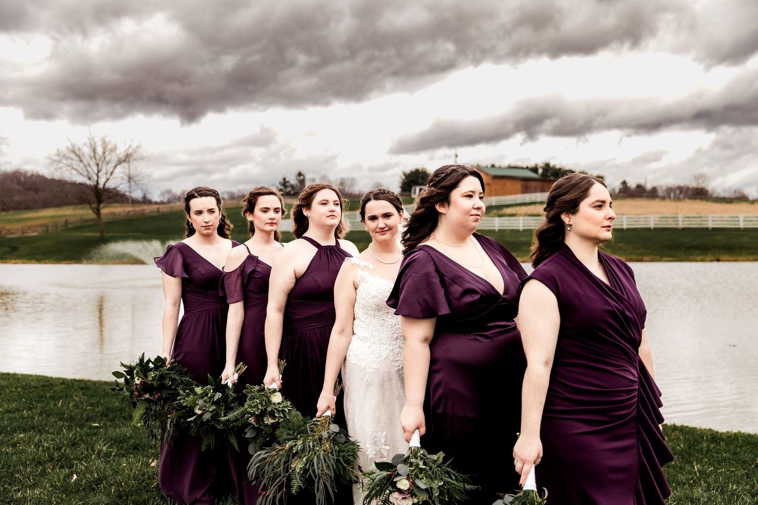 Bride and bridesmaids in deep purple dresses pose by a pond under moody skies at this elegant countryside wedding