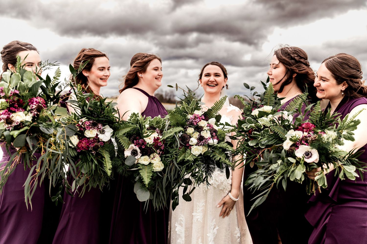 Bride and bridesmaids laugh together holding lush bouquets in this joyful outdoor wedding photo with dramatic skies