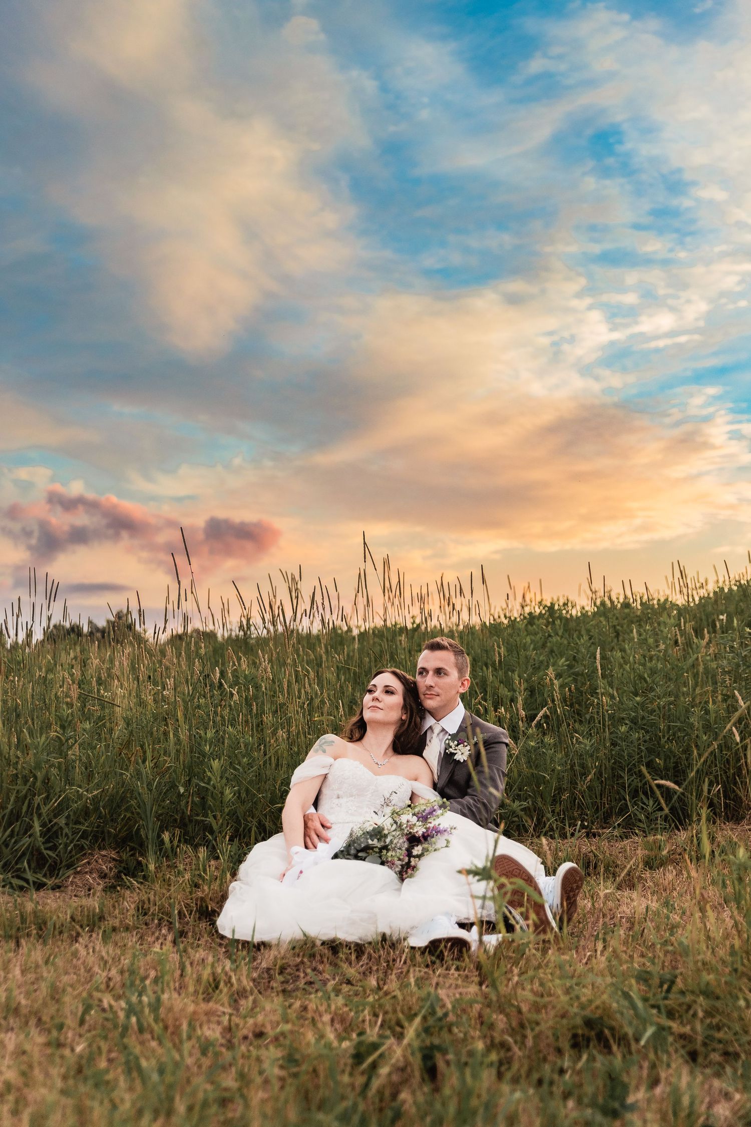 A couple sits in tall grass during sunset with dramatic clouds and warm lighting in the background.
