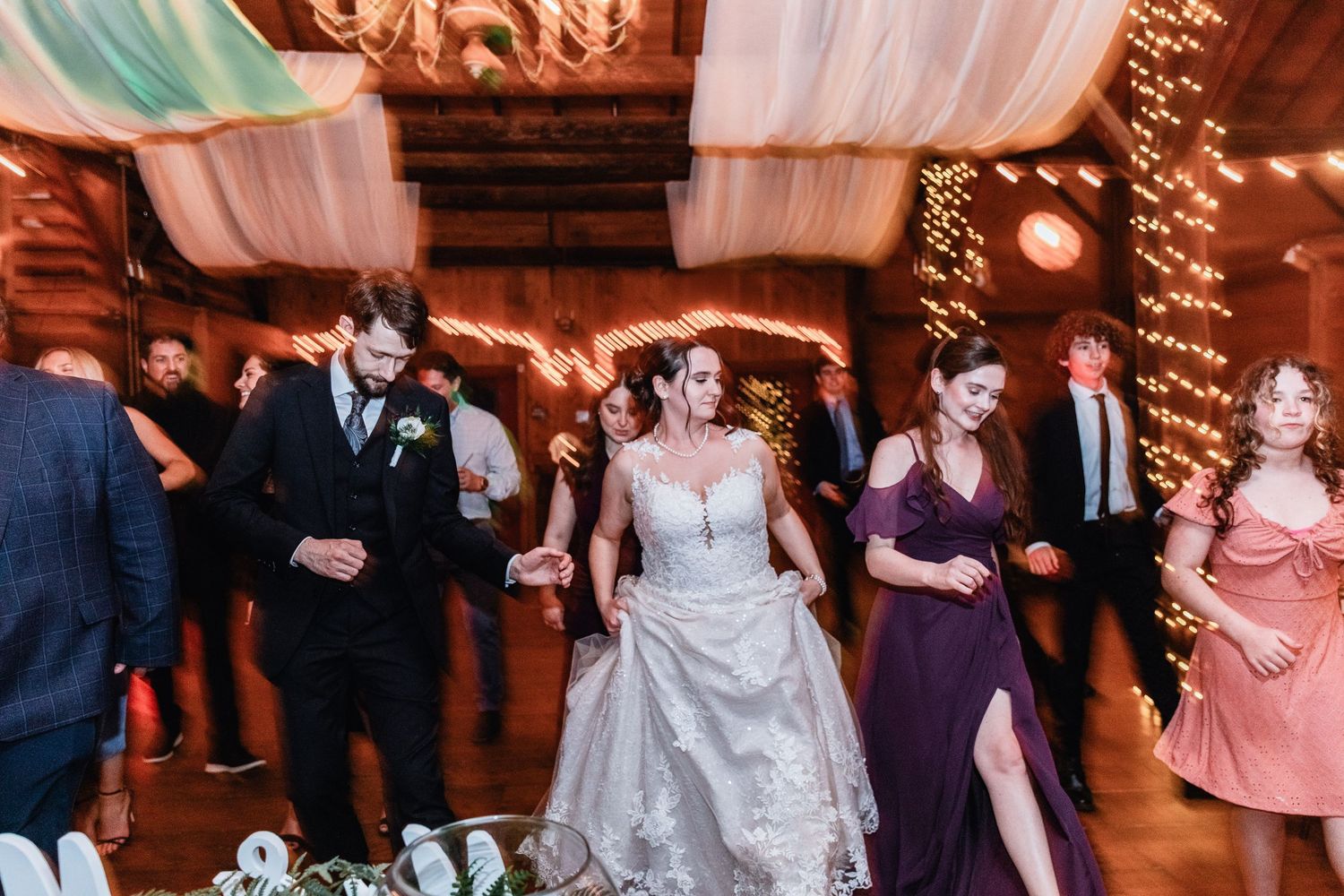 Bride and groom dance with guests at a lively wedding reception in a rustic barn, with twinkle lights and flowing draped fabric