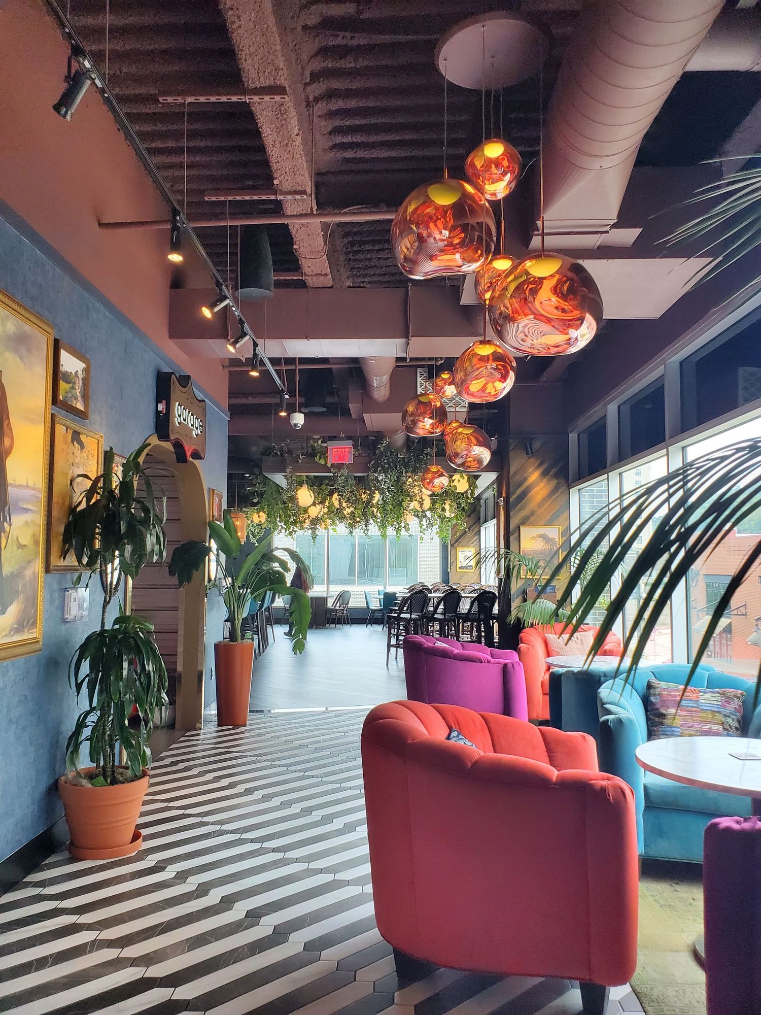 Modern restaurant interior with copper pendant lights, red velvet chairs and striped floors along a windowed wall.