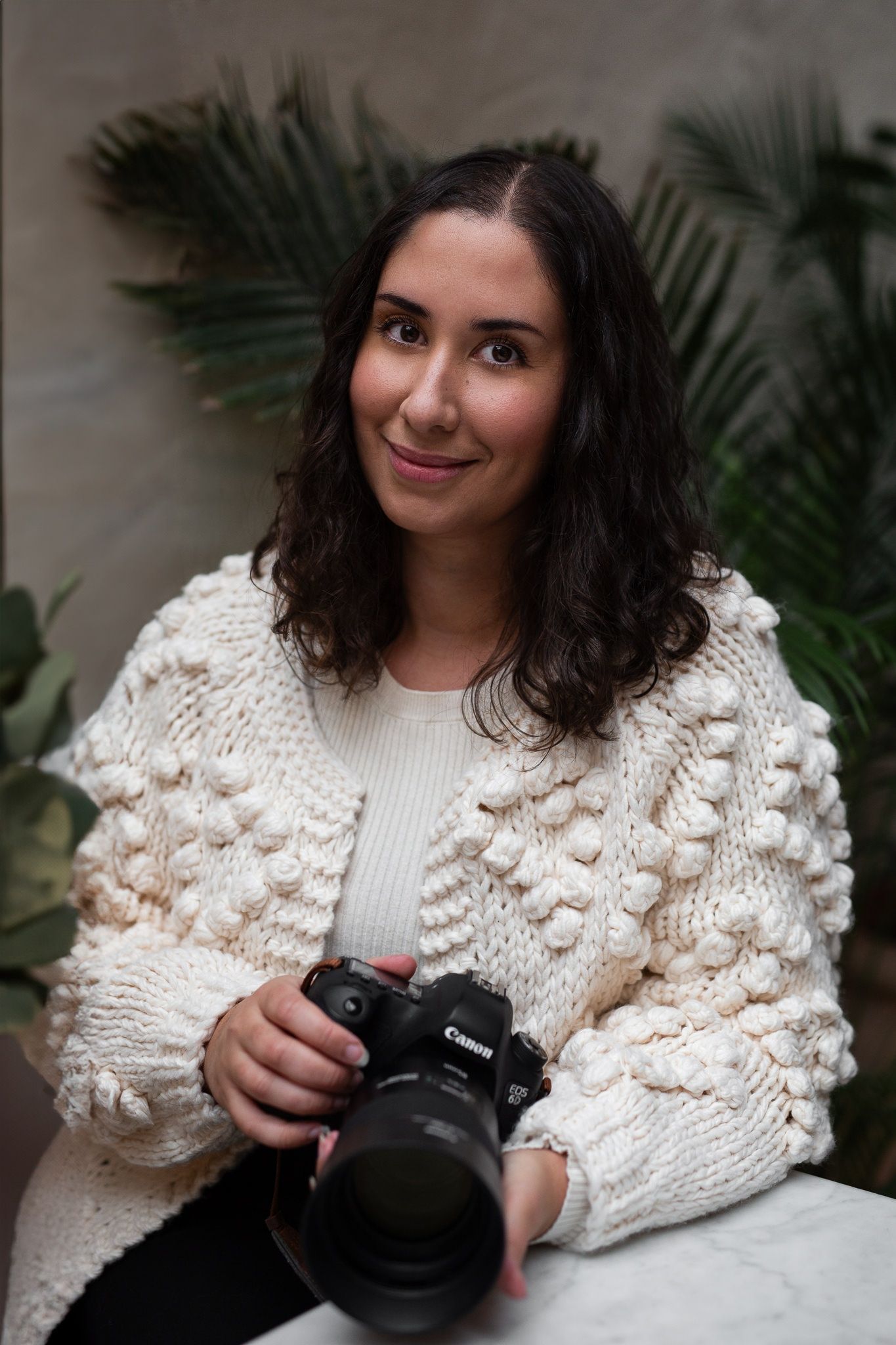 Professional photographer wearing cozy cream knit sweater with pom pom details holds Canon camera in indoor setting with palm leaves.