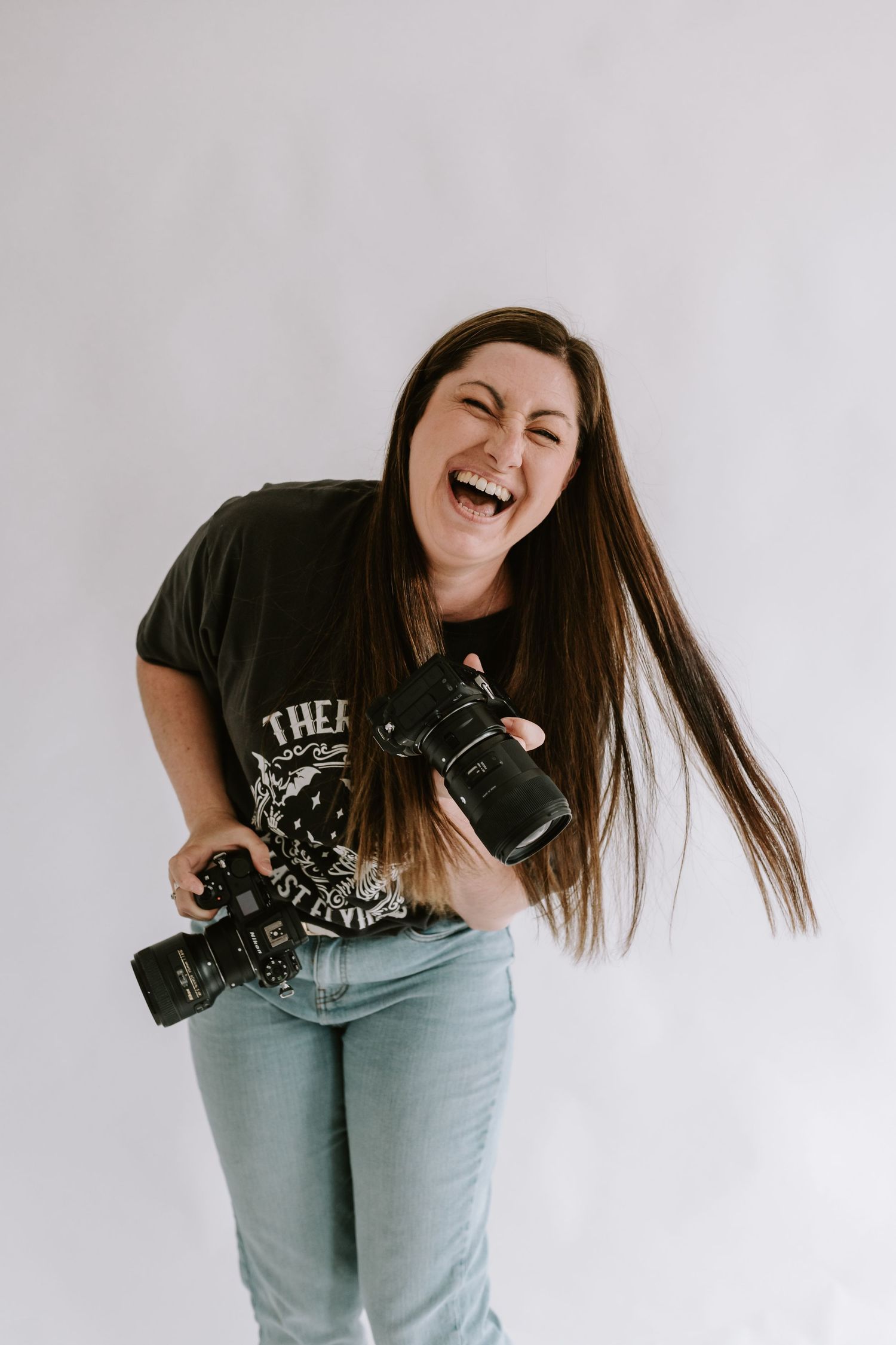 Professional photographer wearing jeans and graphic t-shirt laughs while holding two DSLR cameras against white background.