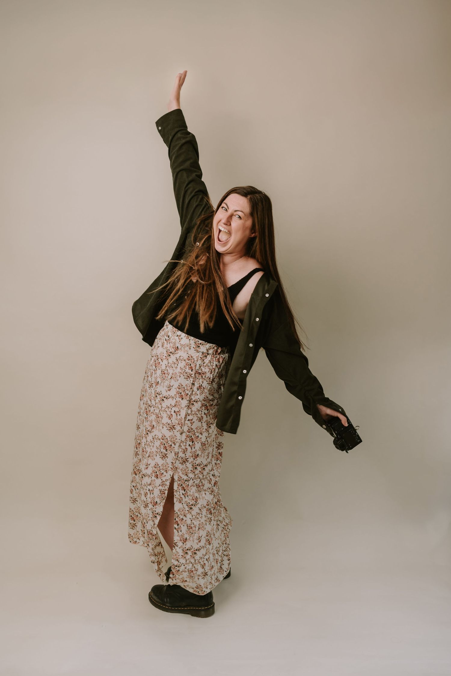 Person in floral dress and dark cardigan strikes playful pose with arm raised against neutral studio backdrop.