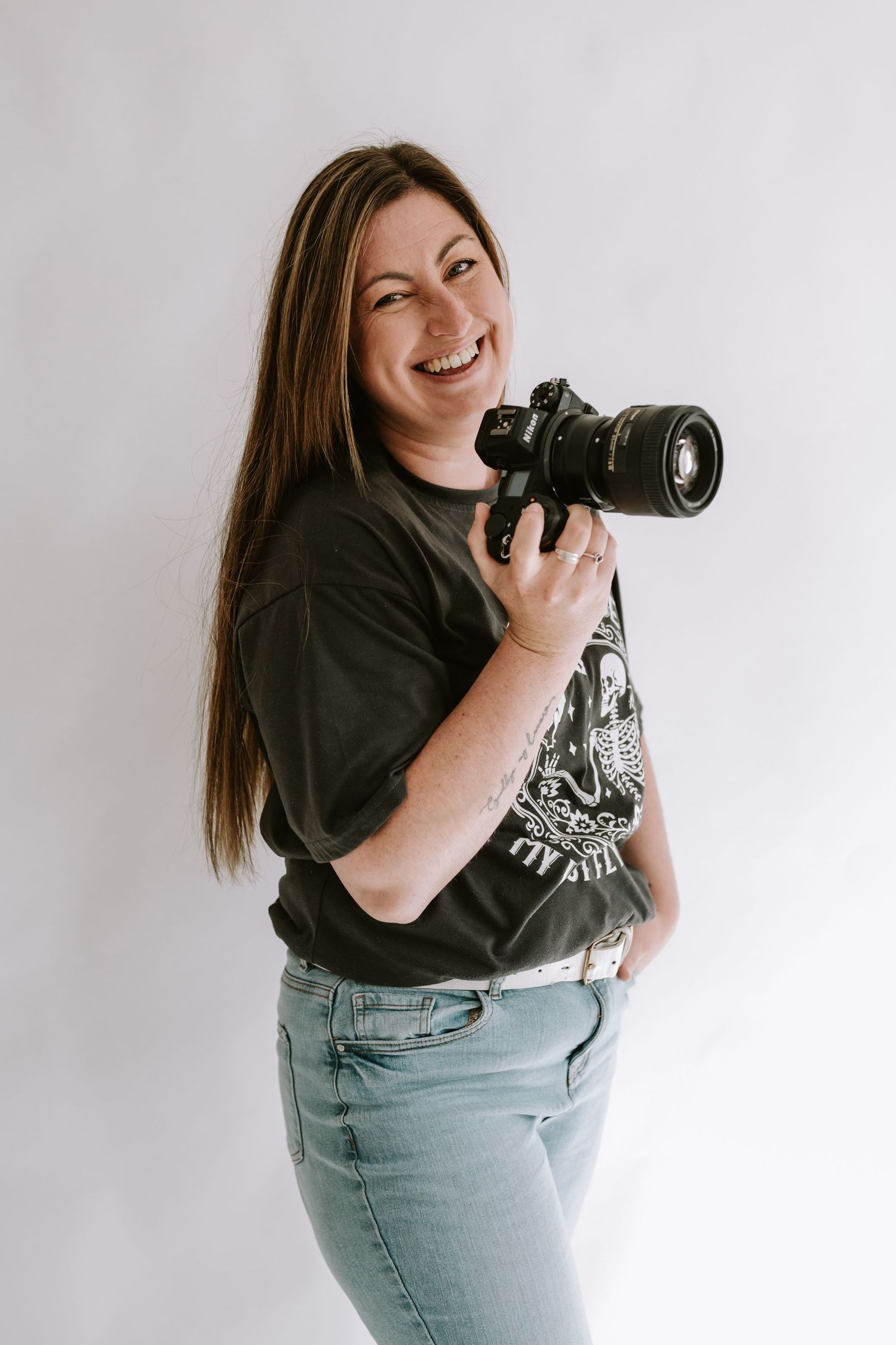 A photographer in a dark t-shirt and jeans holds a professional DSLR camera against a white studio background.