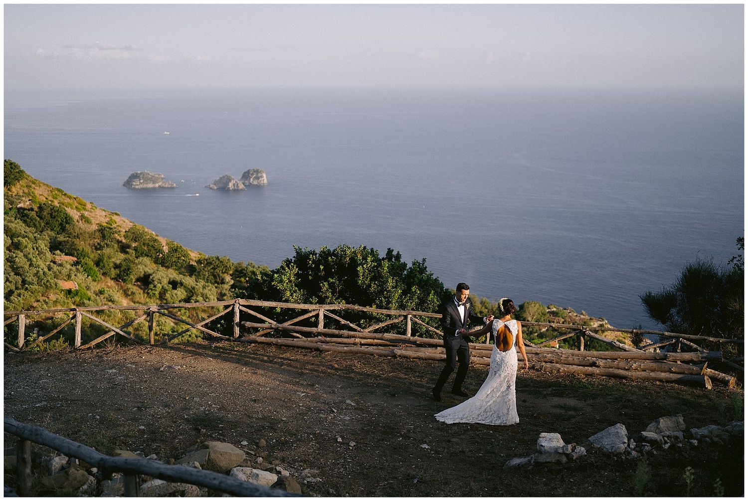 A couple stands at scenic coastal viewpoint with wooden railing overlooking rocky islands in Mediterranean sea at sunset.