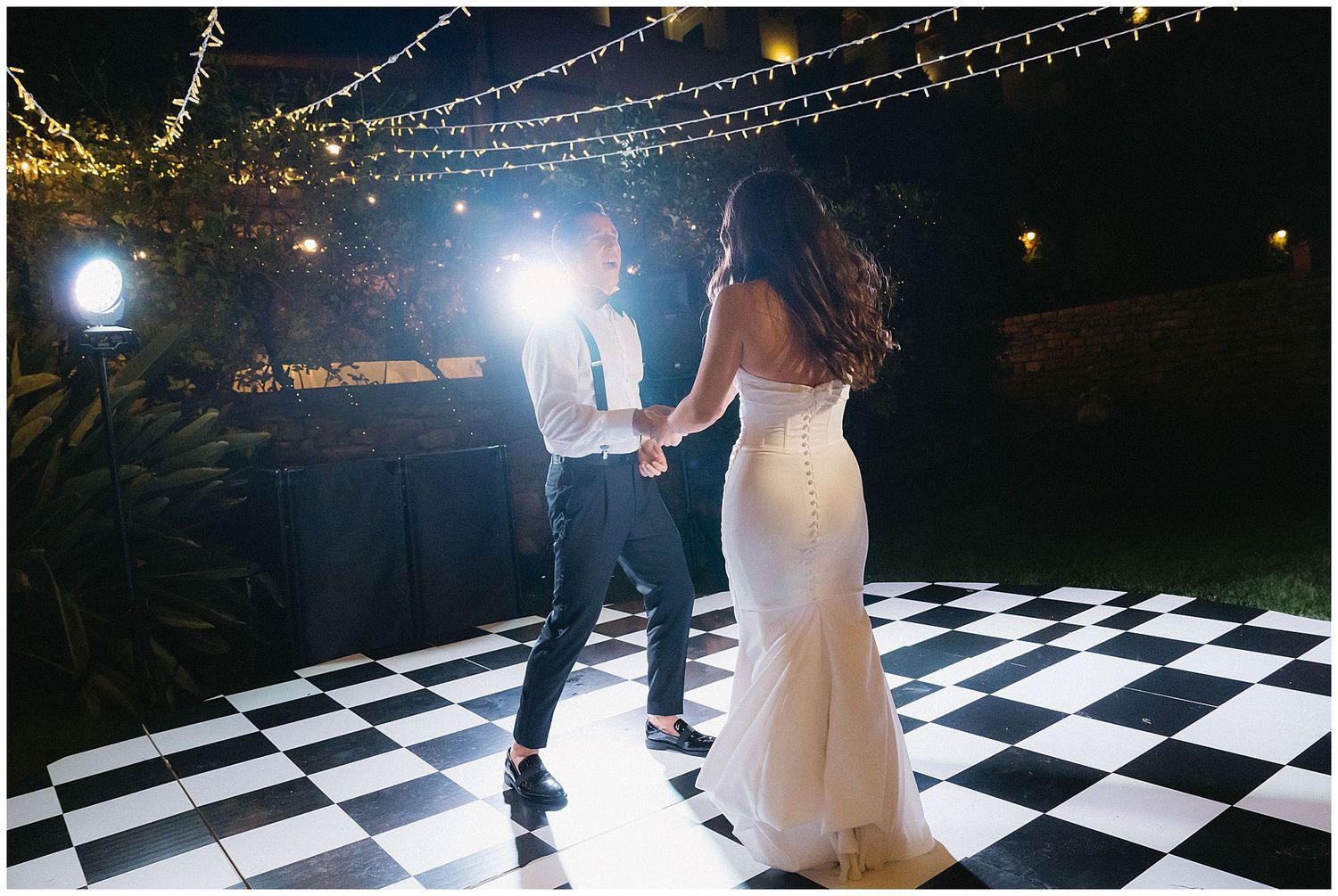 Couple dancing together on black and white checkered dance floor under twinkling lights at evening reception.