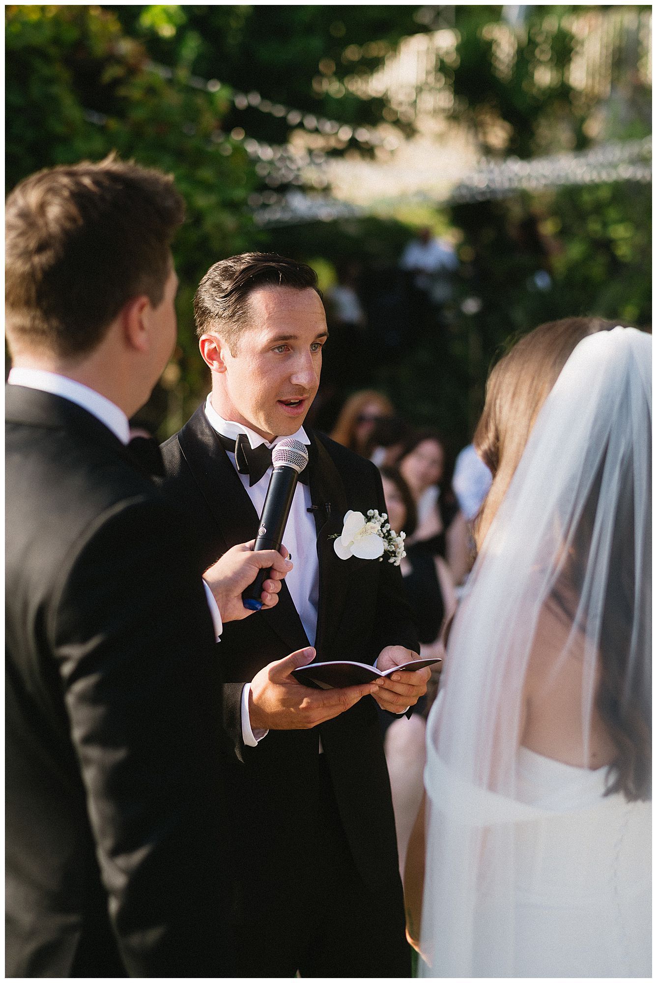 A wedding ceremony in progress shows a groom reading vows while standing with the couple and officiant beneath trees.
