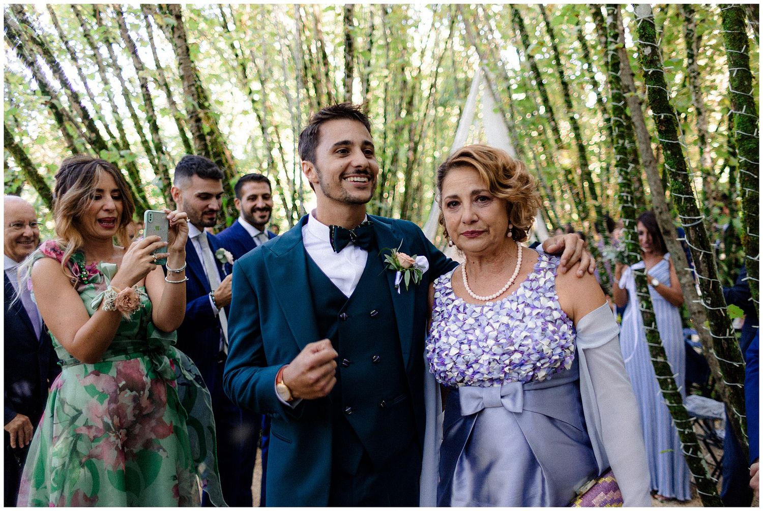 Wedding guests celebrate joyfully with formal attire in an outdoor forest setting while walking down a tree-lined path.