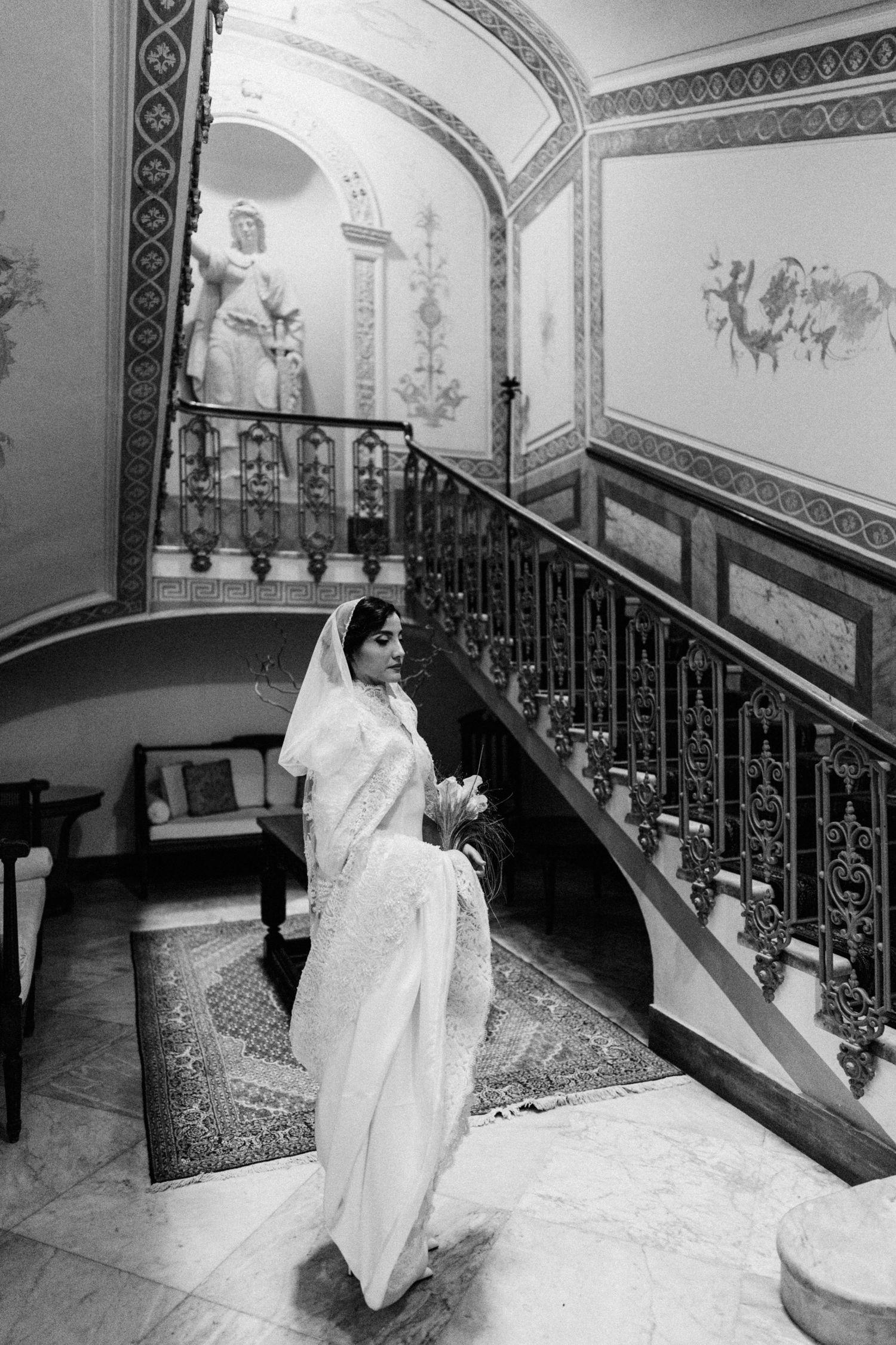 A person in a white sari stands elegantly on a grand marble staircase with ornate railings and decorative wall murals.