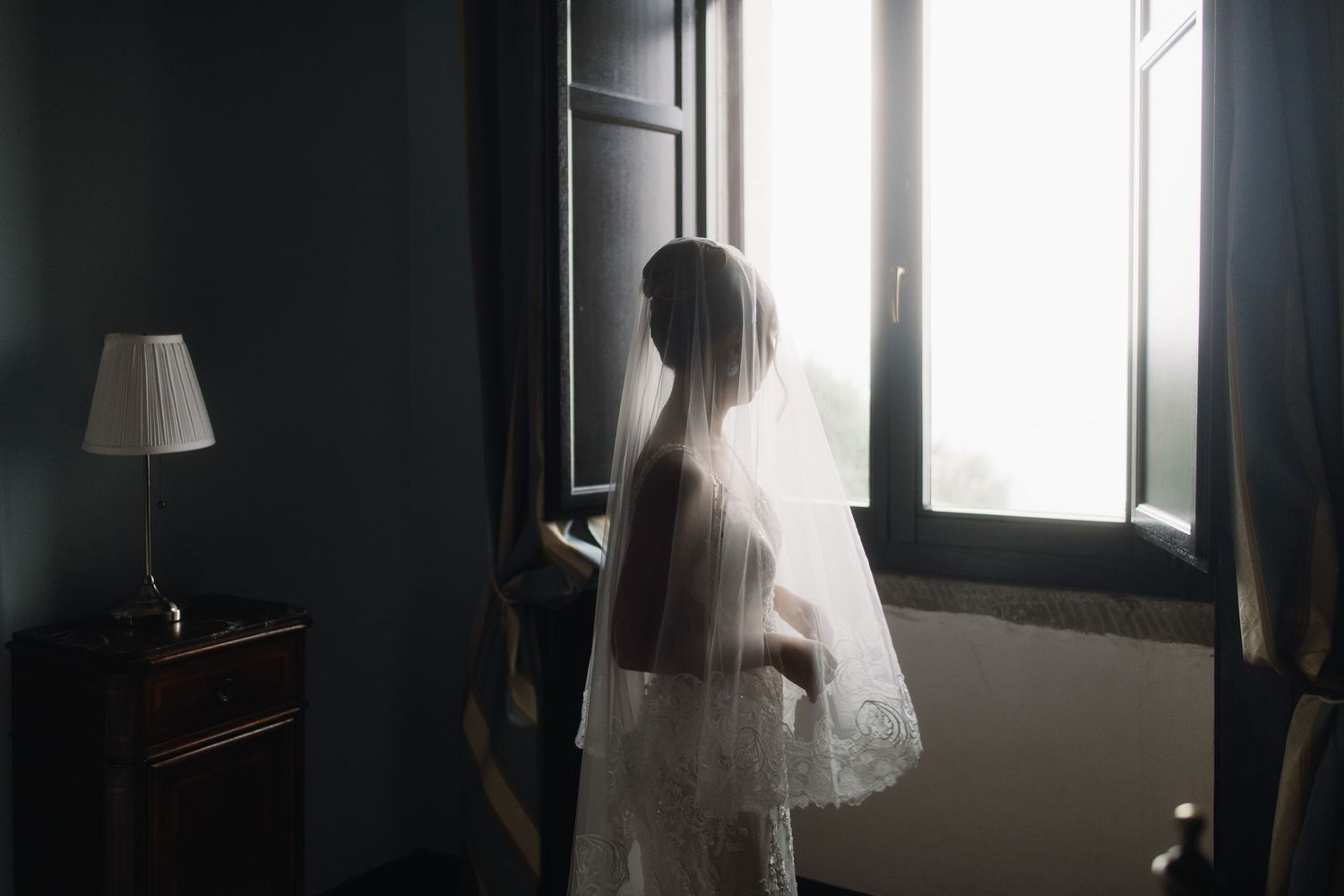 A person in a flowing white wedding veil gazes out a window in a dimly lit room with a table lamp nearby.