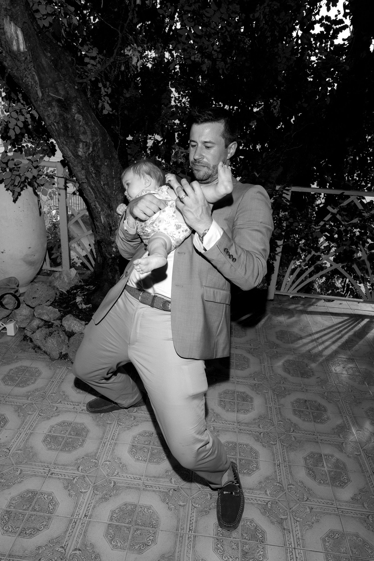 Someone in a grey suit dances playfully while holding a small baby at an outdoor event.
