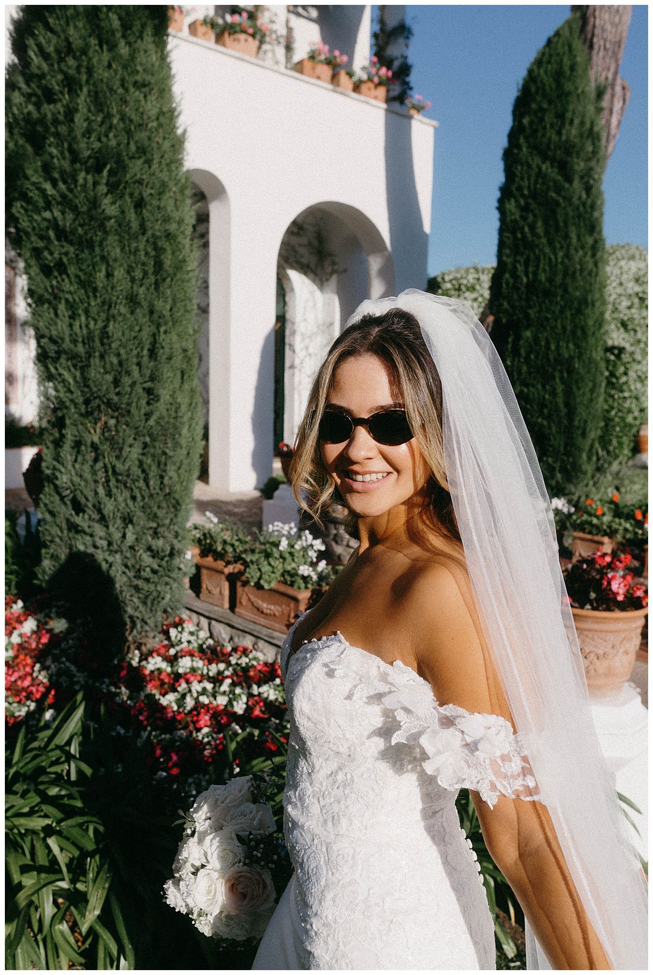 A bride wearing a white off-shoulder lace gown and sunglasses poses by cypress trees and white arches on her wedding day.