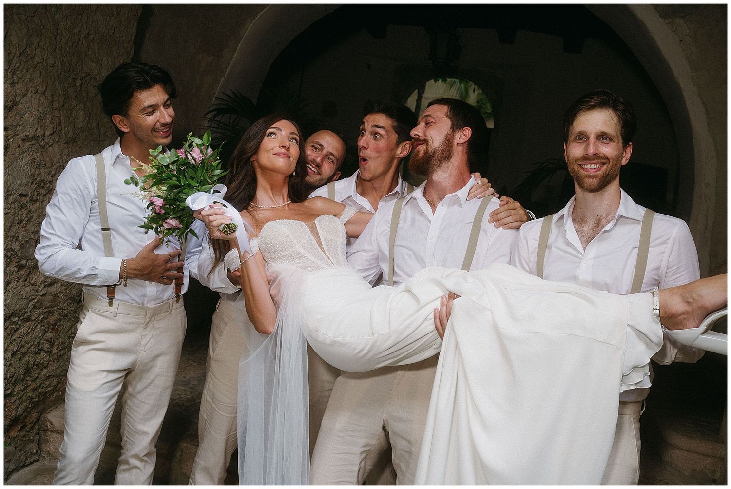 A joyful wedding party in white attire shares a candid laughing moment under a stone archway.