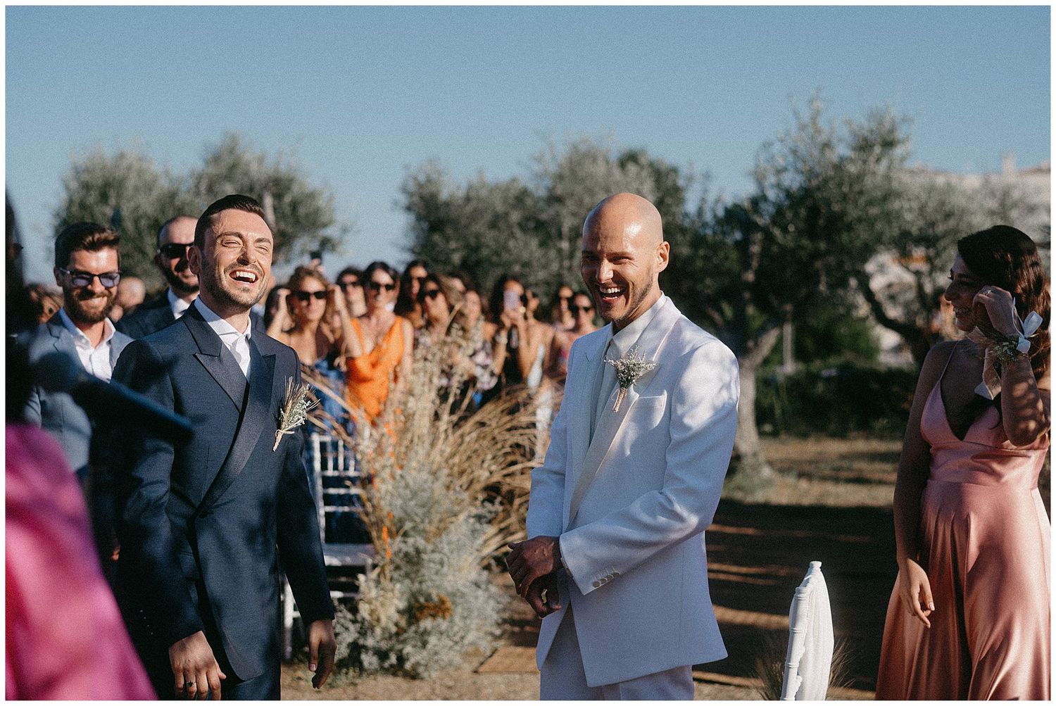 Two people sharing a joyful moment at an outdoor wedding ceremony with guests and olive trees in the background.
