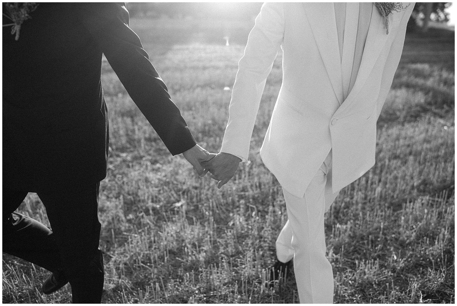 Black and white close-up of couple holding hands in a grassy field with sun flare creating romantic ambiance.