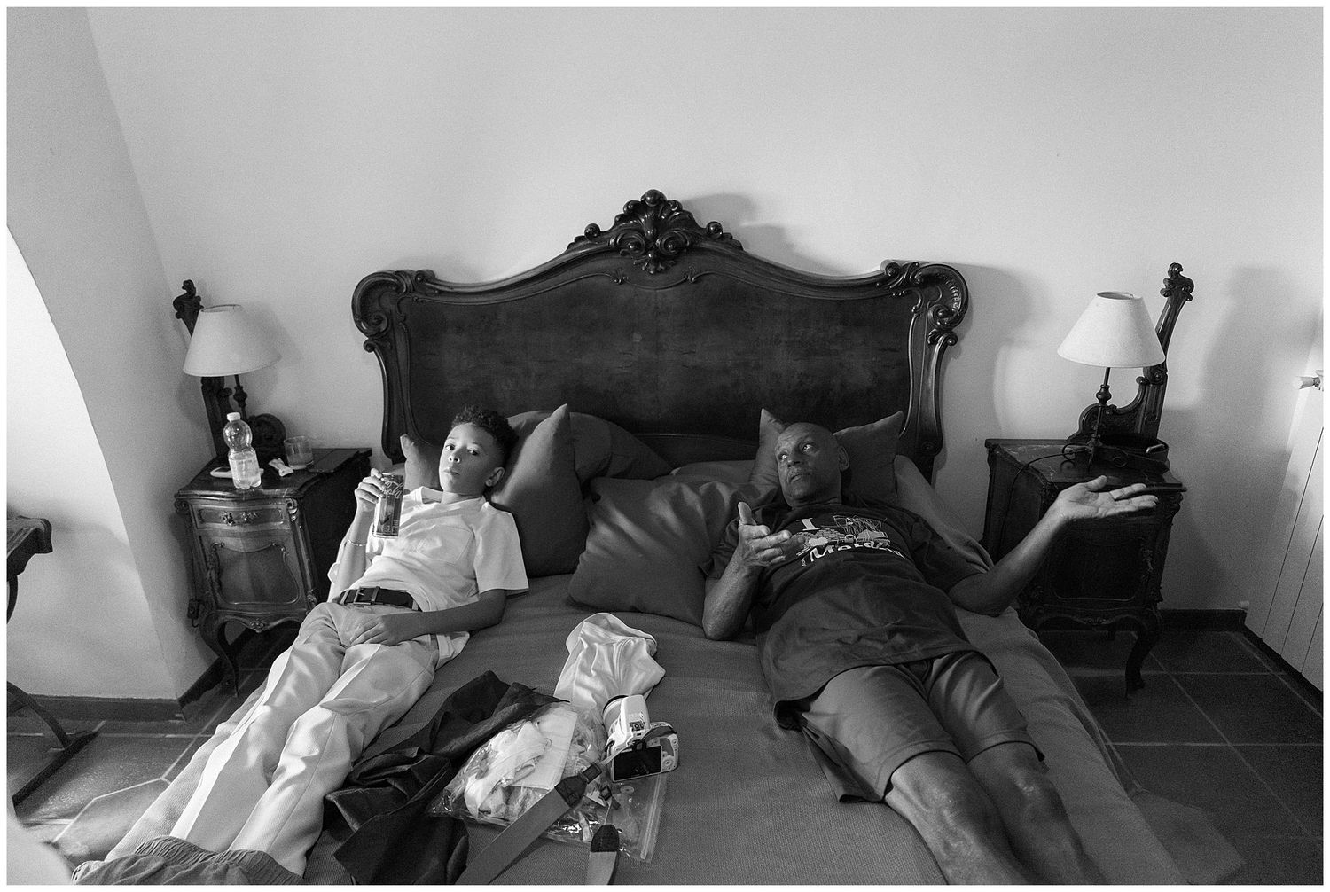 Black and white photograph of two people lying on a bed with an ornate headboard in a domestic setting.