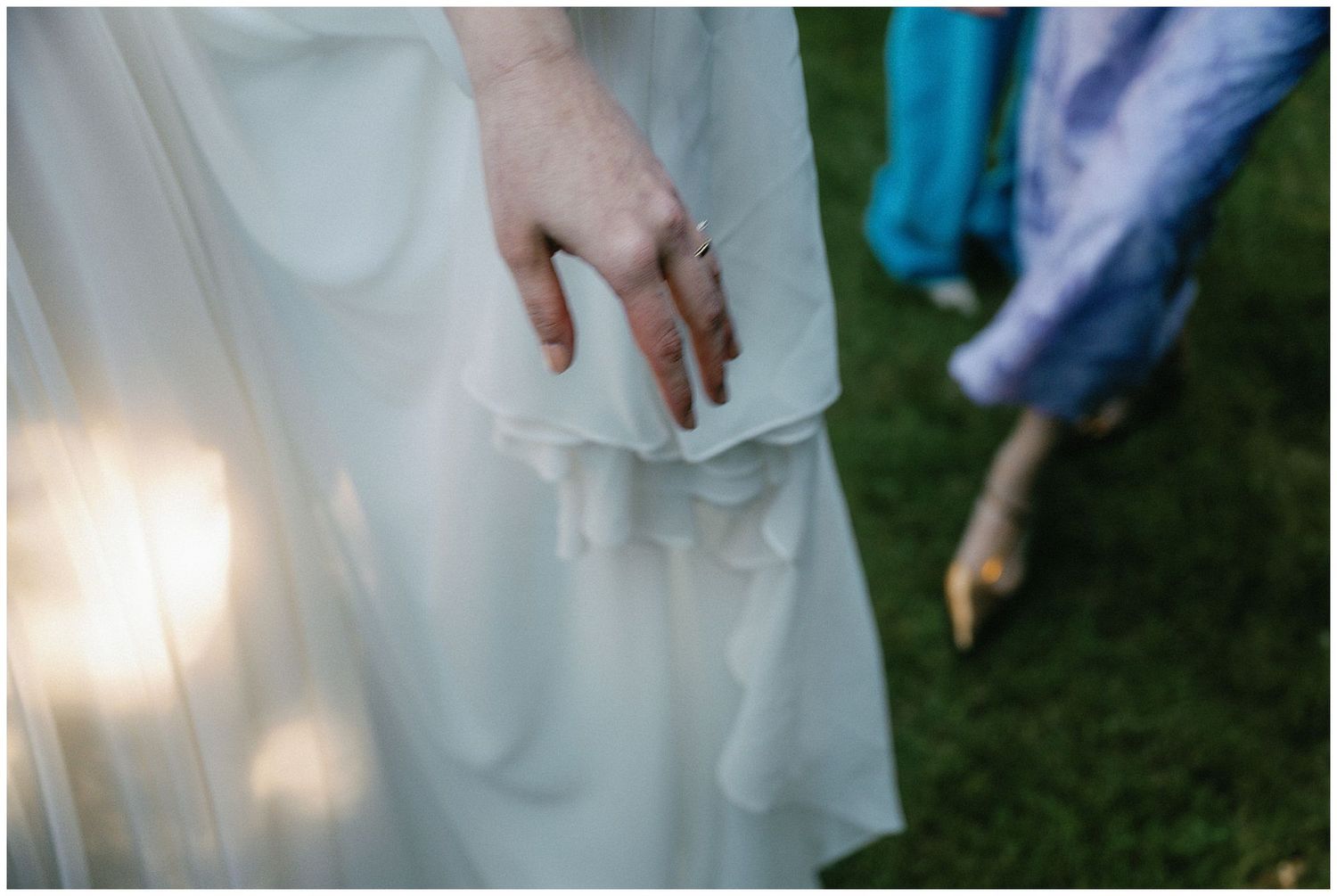 Close up detail of a hand delicately holding white fabric of a dress against a grassy outdoor background.