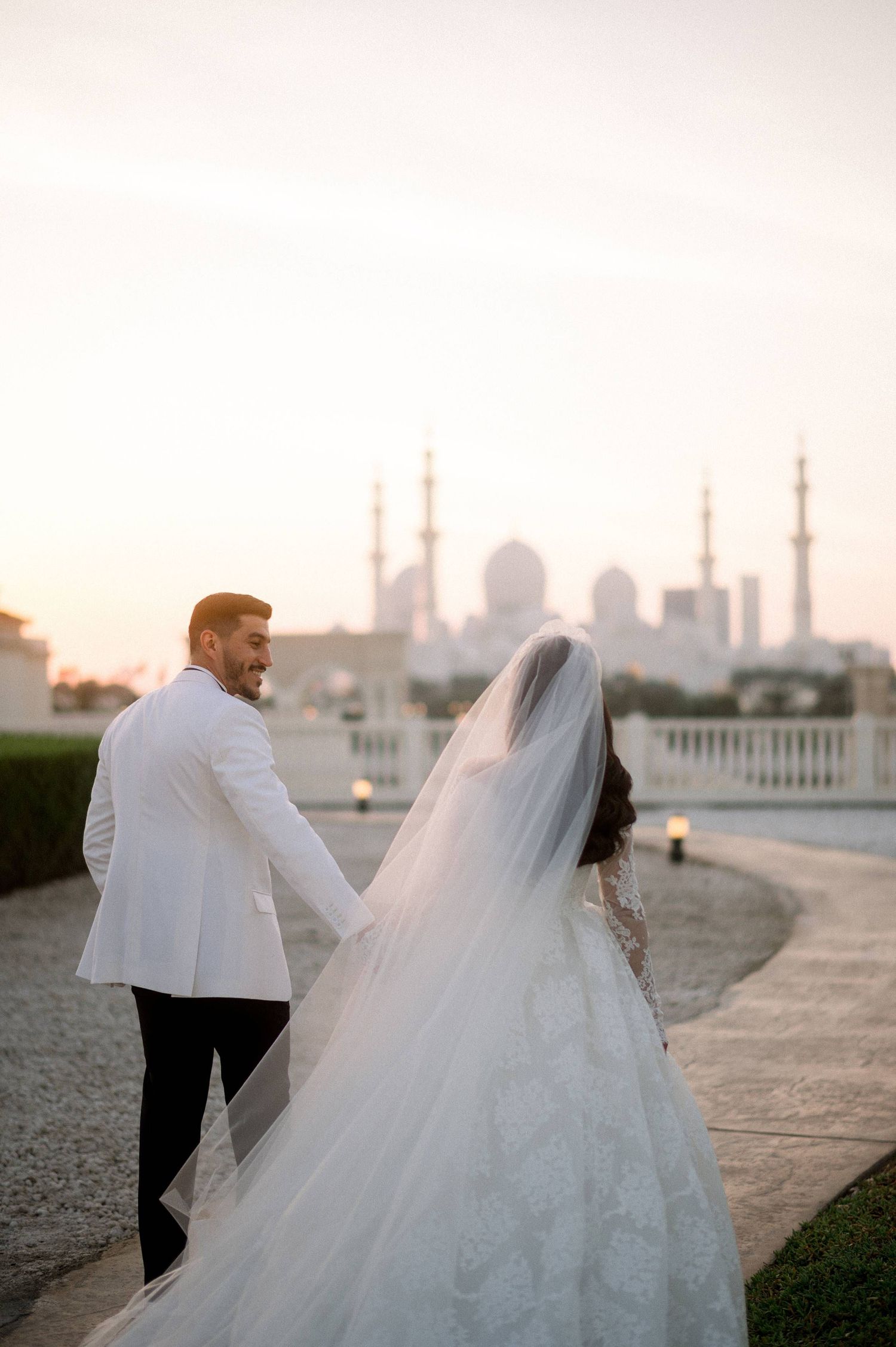 A couple in wedding attire holds hands overlooking the Sheikh Zayed Grand Mosque in Abu Dhabi during sunset.
