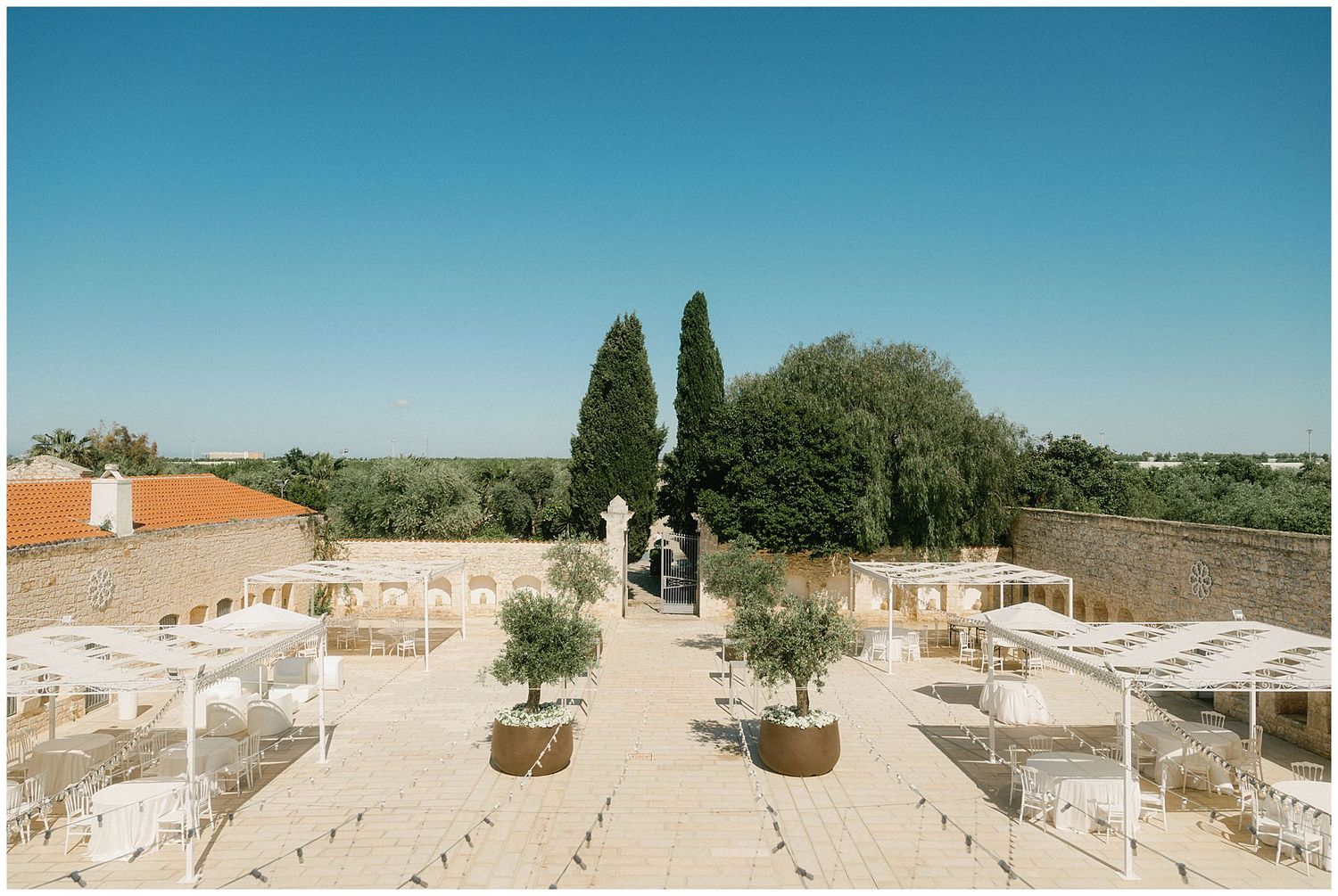 People strolling through a rooftop garden terrace with cypress trees and white walls overlooking a sunny Mediterranean landscape.