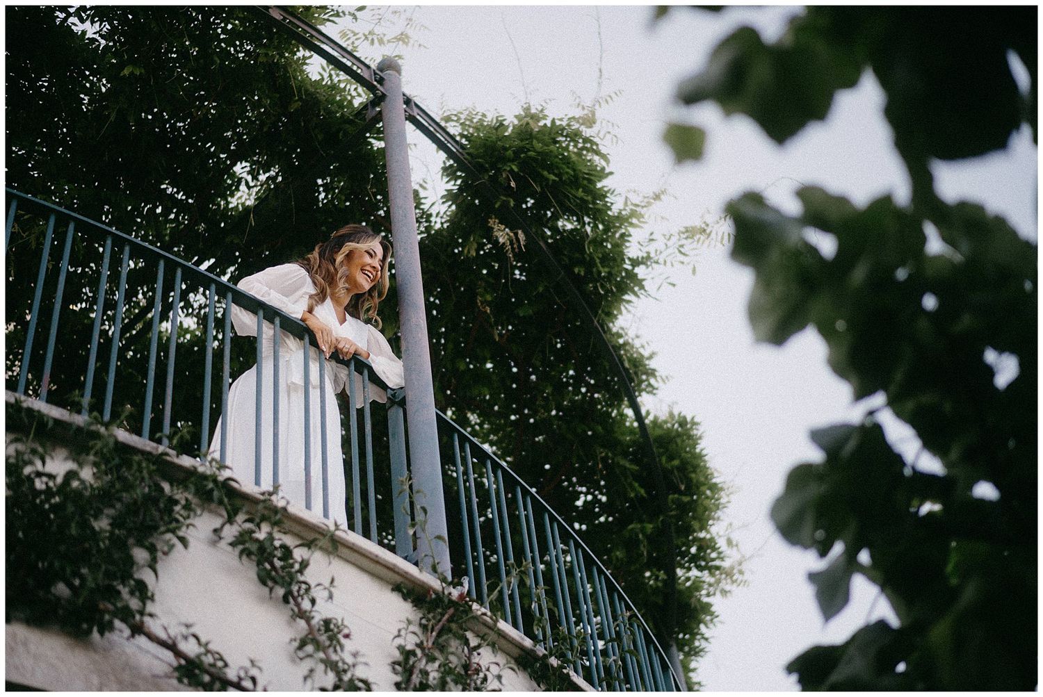 A figure leans over a turquoise wrought iron balcony railing on a white building surrounded by lush green trees.