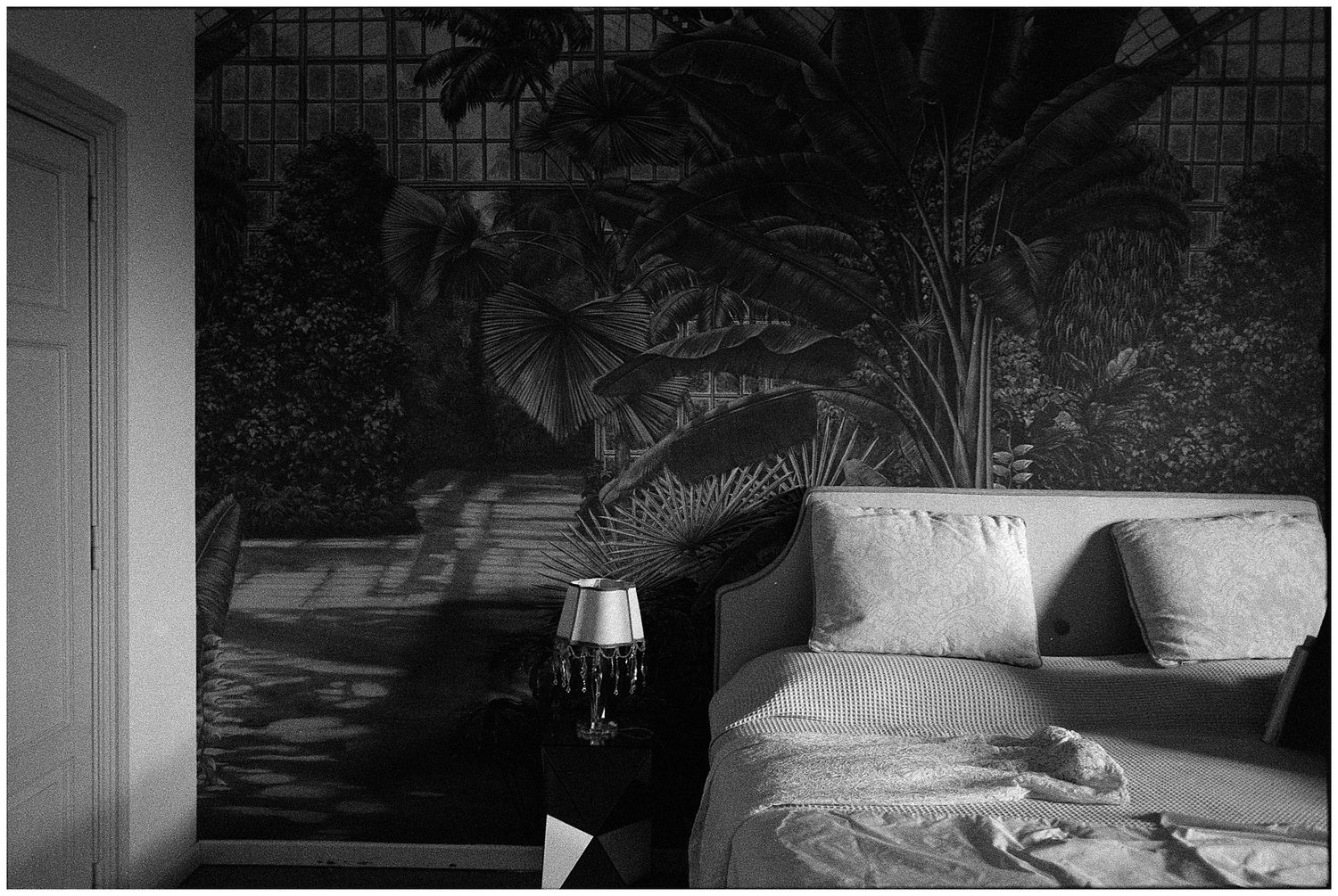 Black and white interior photo showing a bed with white linens against a dark floral wallpapered wall with soft lighting.