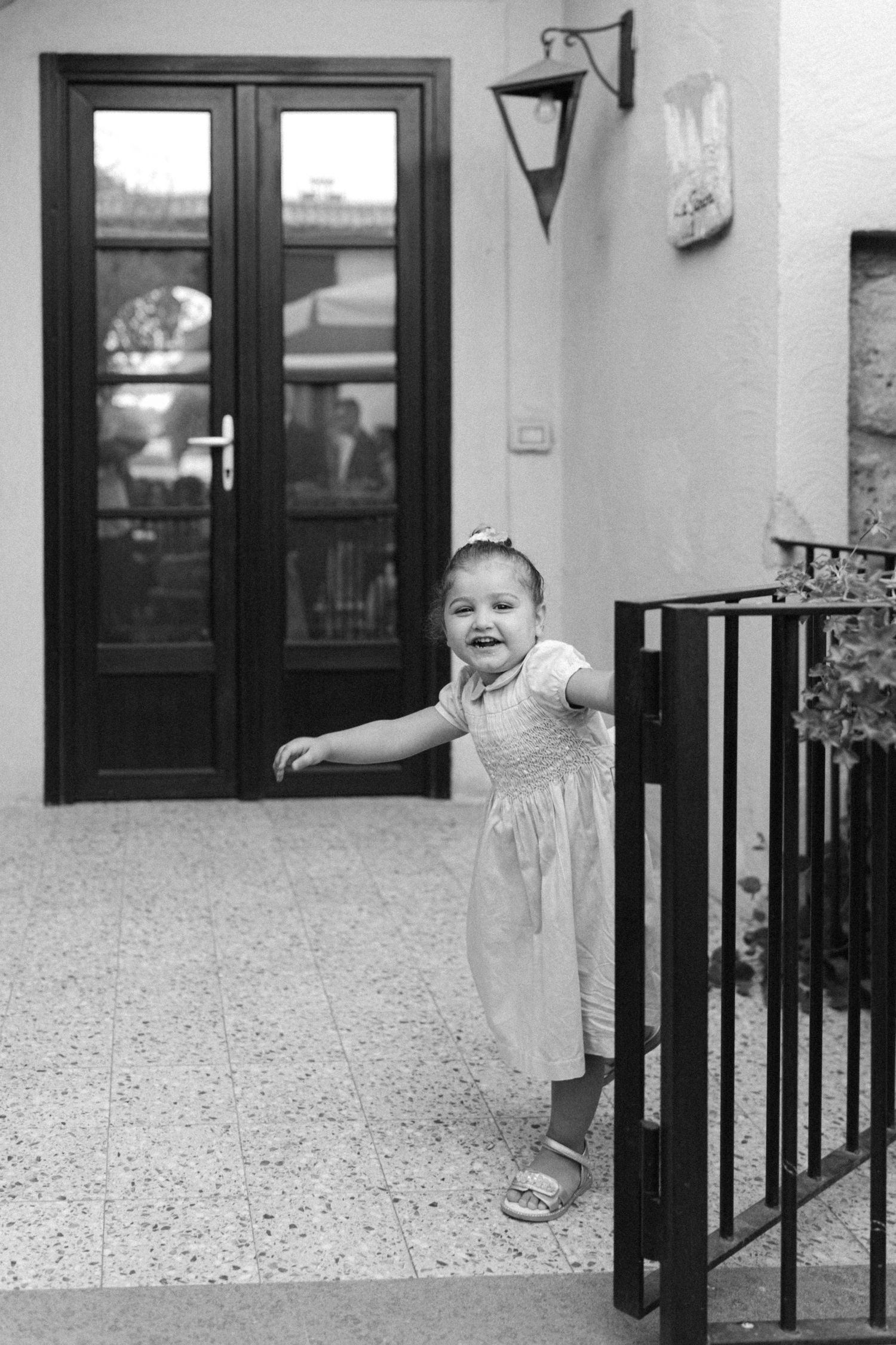 Black and white photo of a toddler in a white dress standing by a railing near glass doors.
