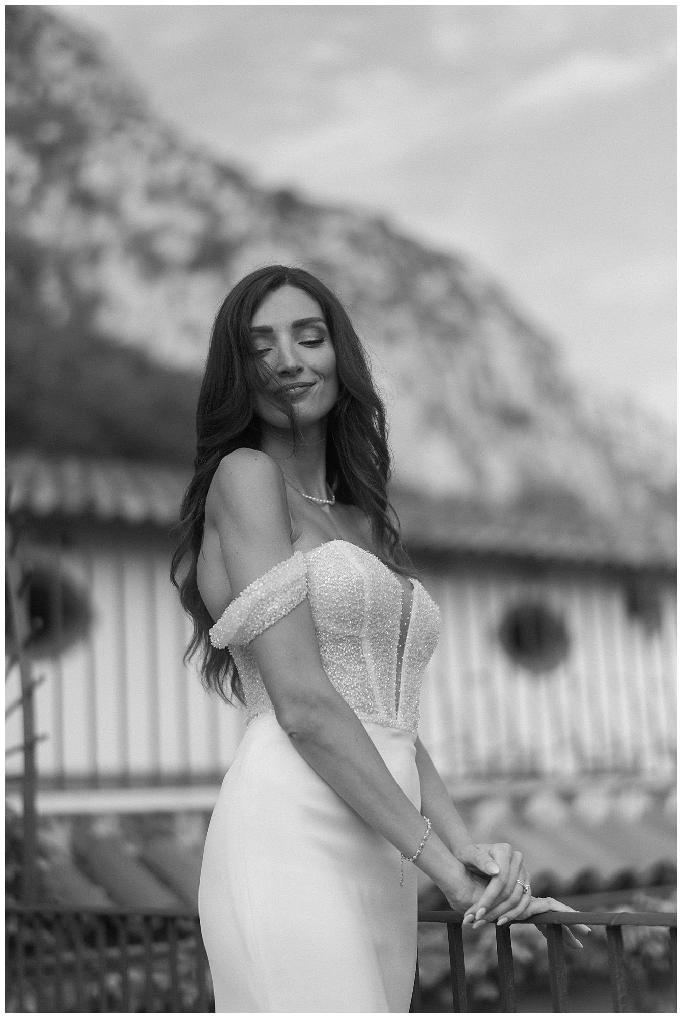 Black and white portrait of a person in an elegant off-shoulder wedding dress posing outdoors against a mountain backdrop.