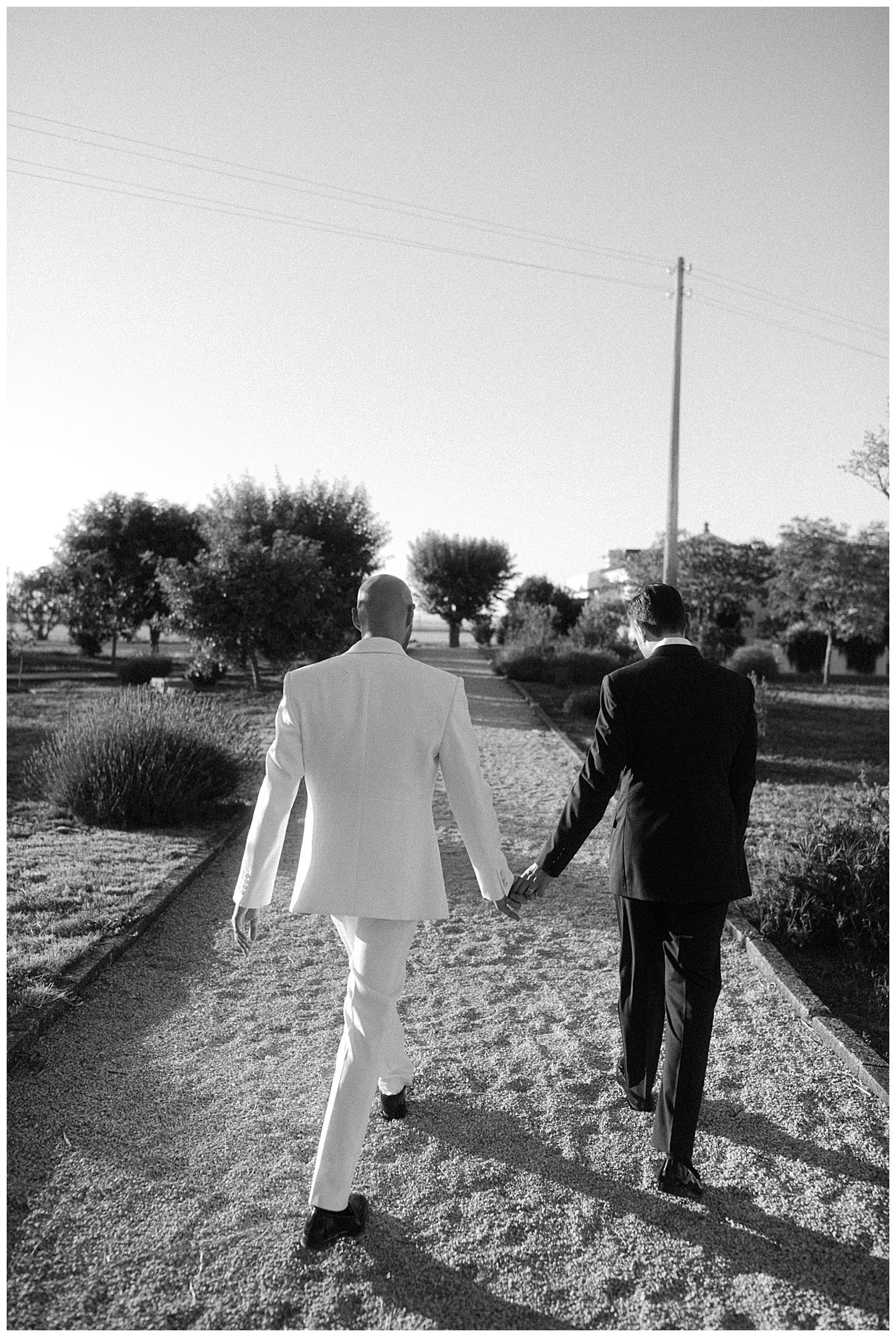 Two people in contrasting black and white suits walking together on a garden path in a black and white photograph.