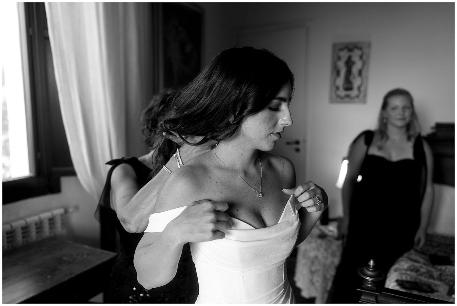 A black and white photograph of someone getting dressed in a white wedding gown with another person in the background.