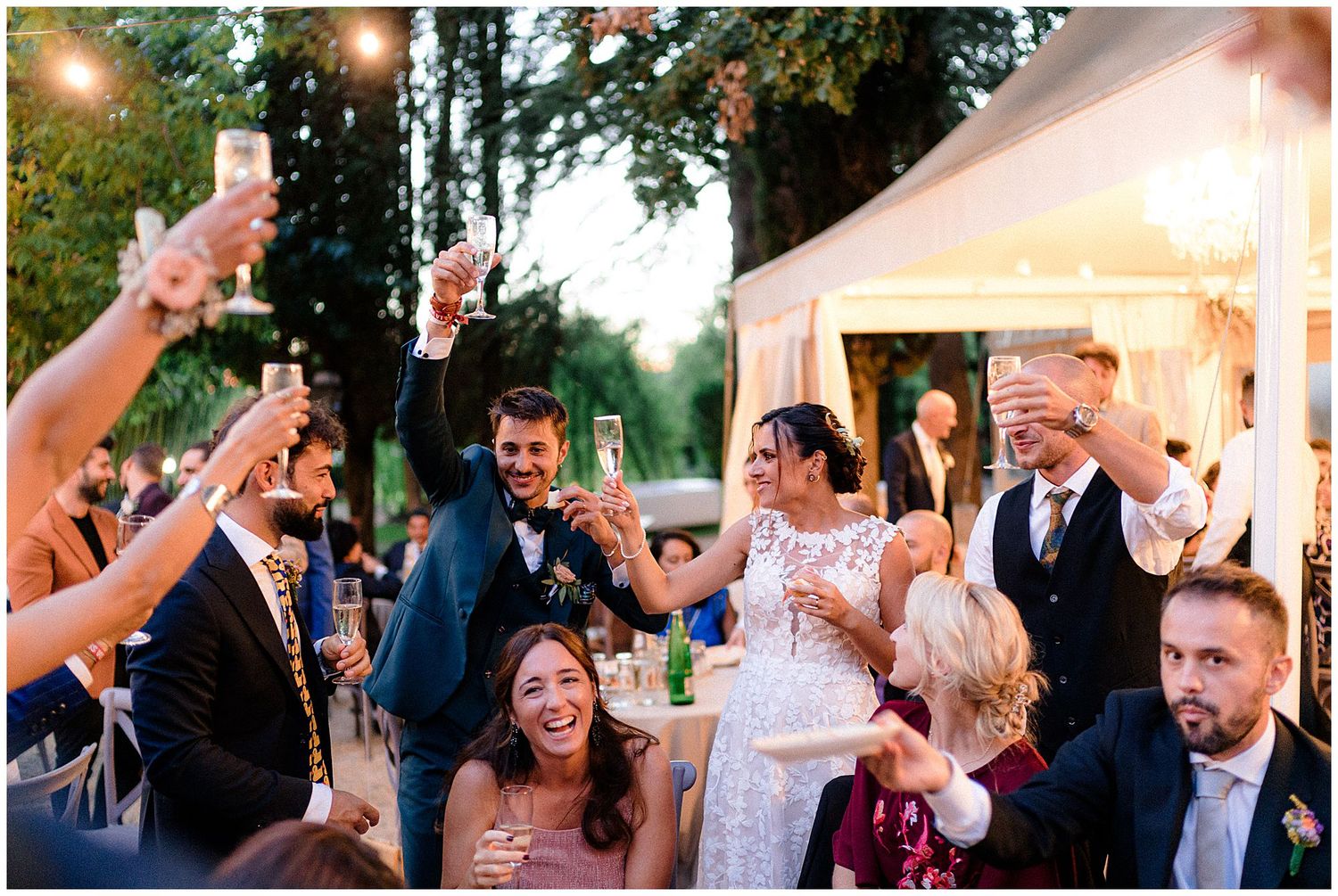 Outdoor wedding celebration with guests toasting under a white tent at dusk.