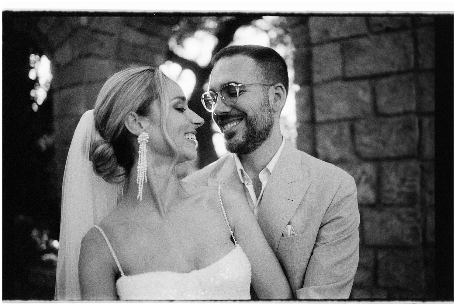 A joyful newlywed couple share a candid moment against a stone wall background during their black and white wedding photoshoot.