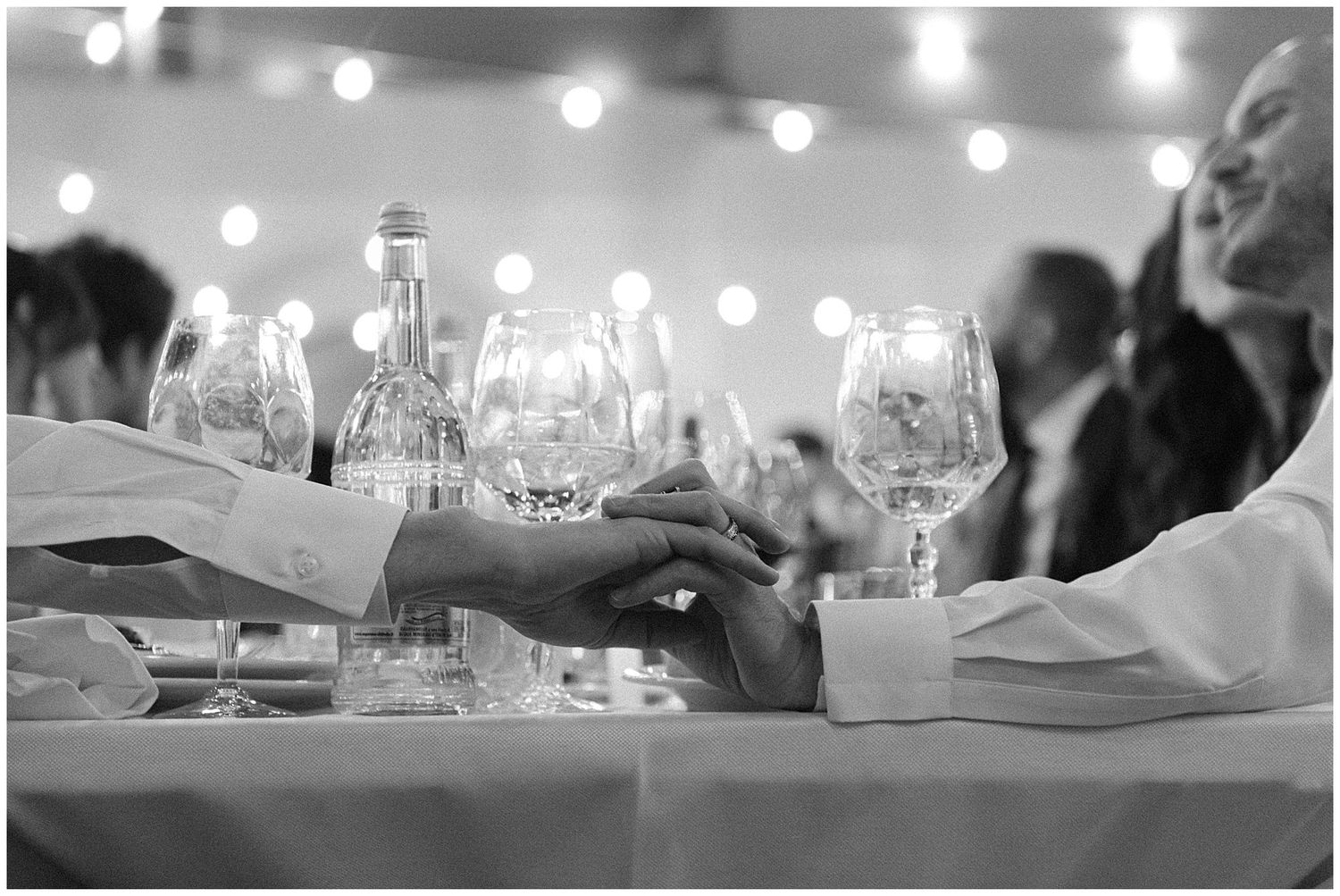 Black and white image of wine glasses and place settings on a table with twinkle lights in the background.