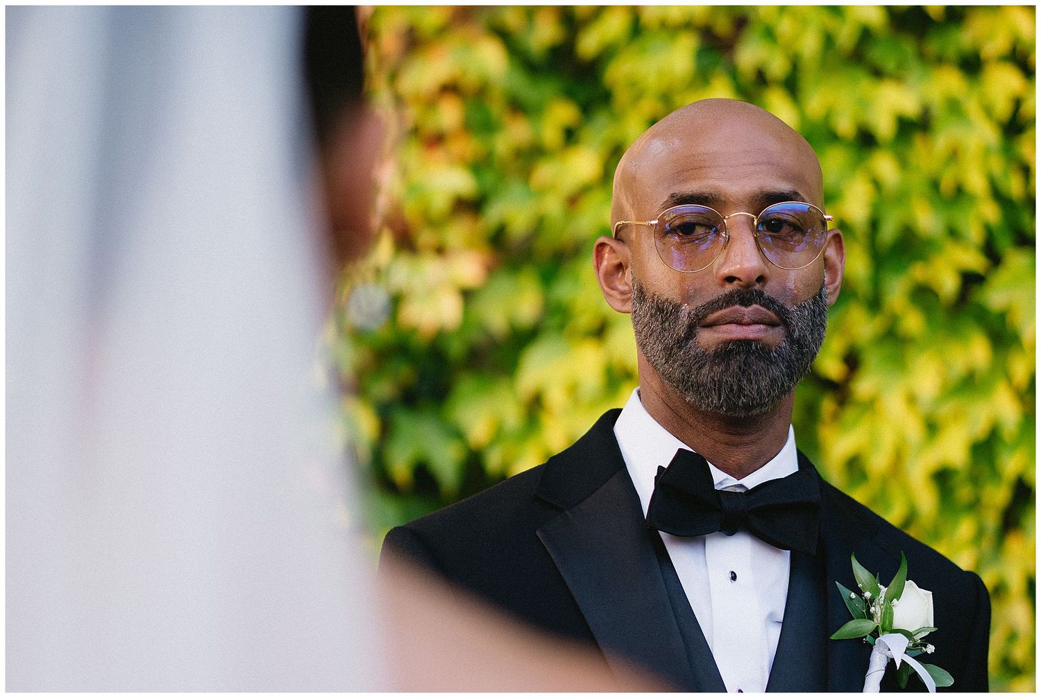 A wedding ceremony image showing a groom in a black tuxedo with a white boutonniere against a green ivy backdrop.