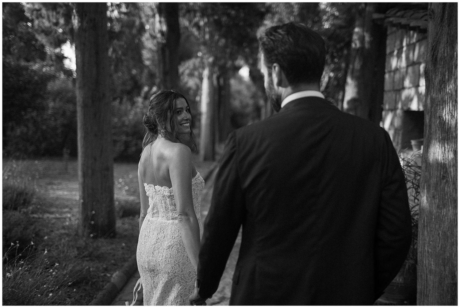 Black and white photo of a couple in wedding attire holding hands while walking through a wooded area.