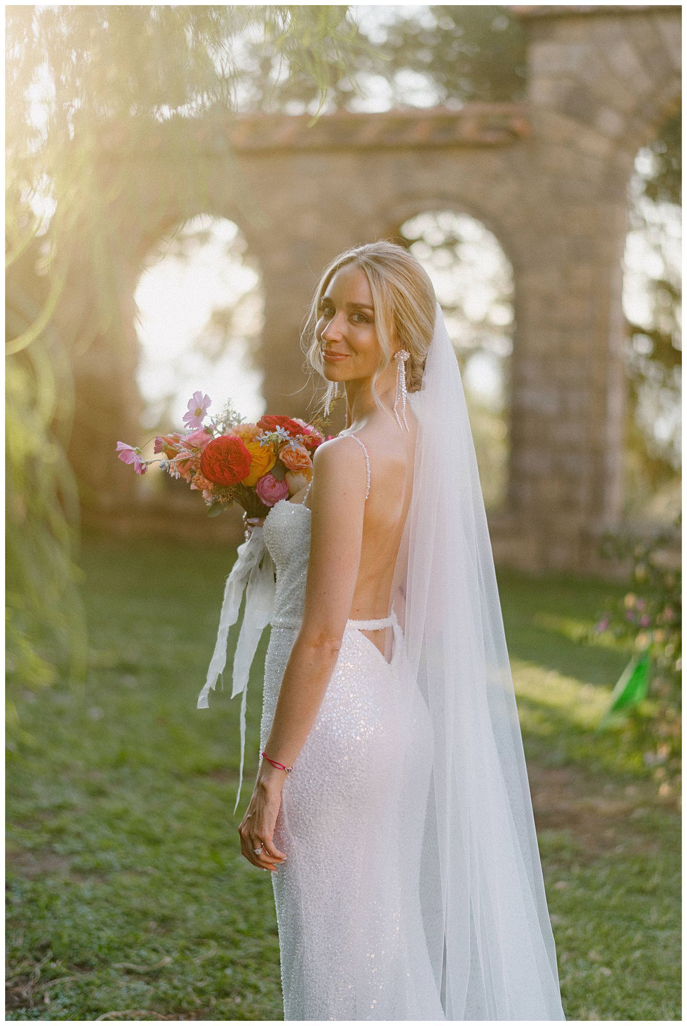 A bride in a flowing white dress and veil holds a colorful bouquet in front of stone archways outdoors.