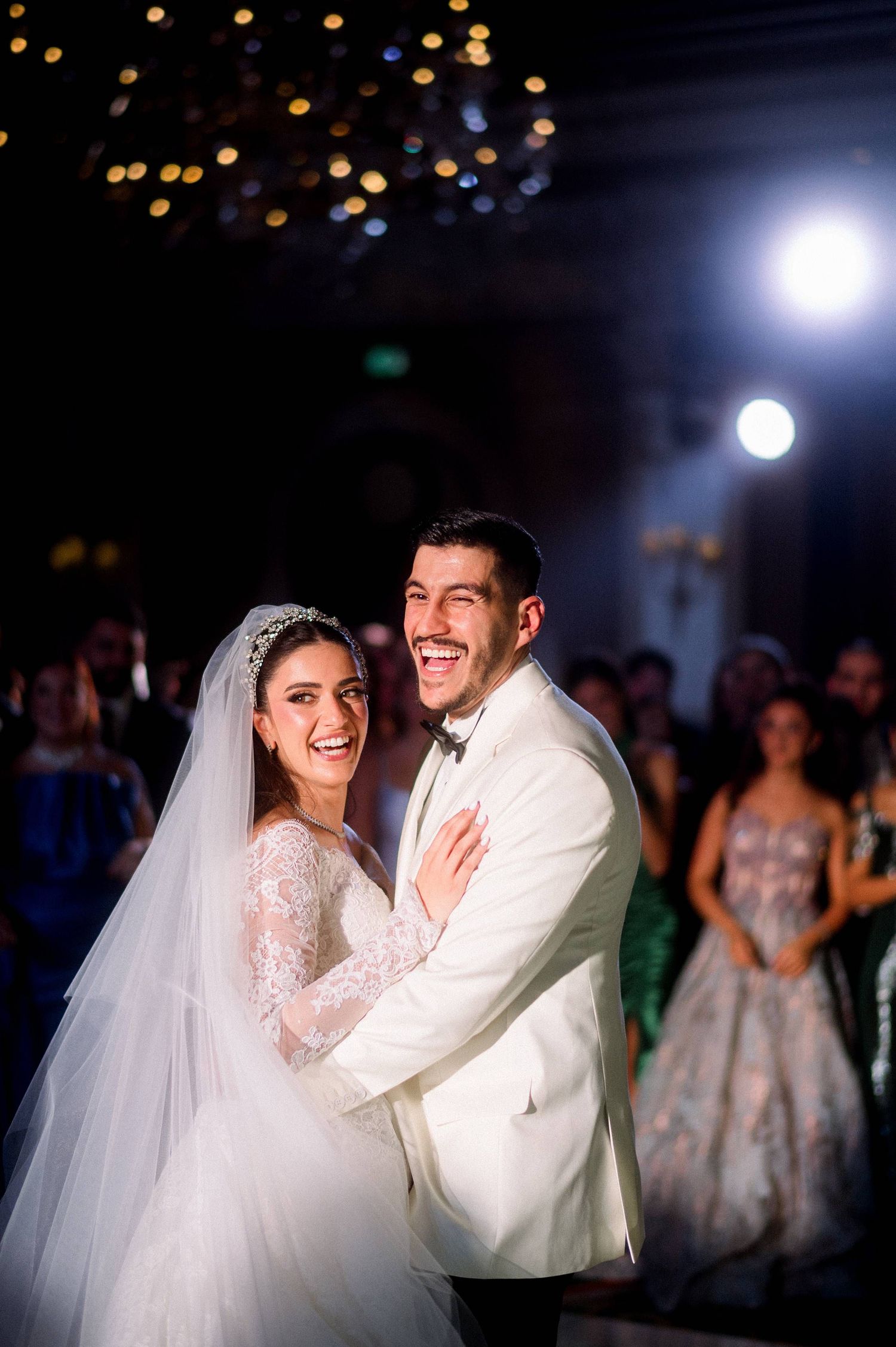 A wedding couple shares joyful laughter while dancing together at their reception celebration under dramatic lighting.