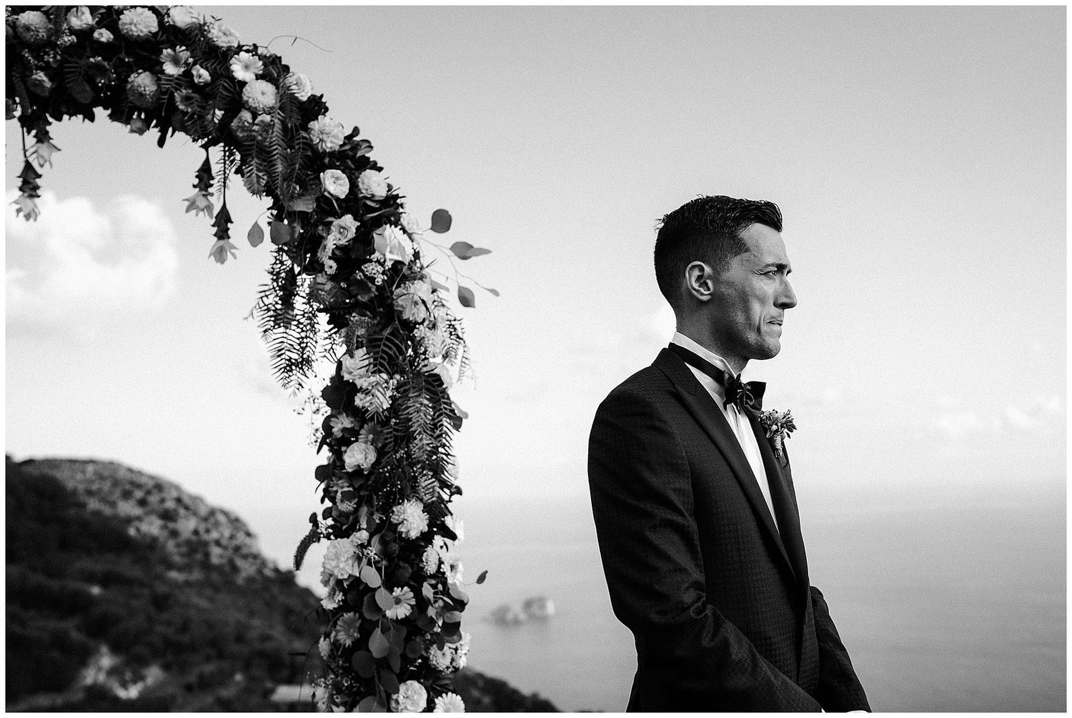 Black and white wedding photo featuring a tuxedo-clad groom standing beside a floral arch overlooking the ocean.