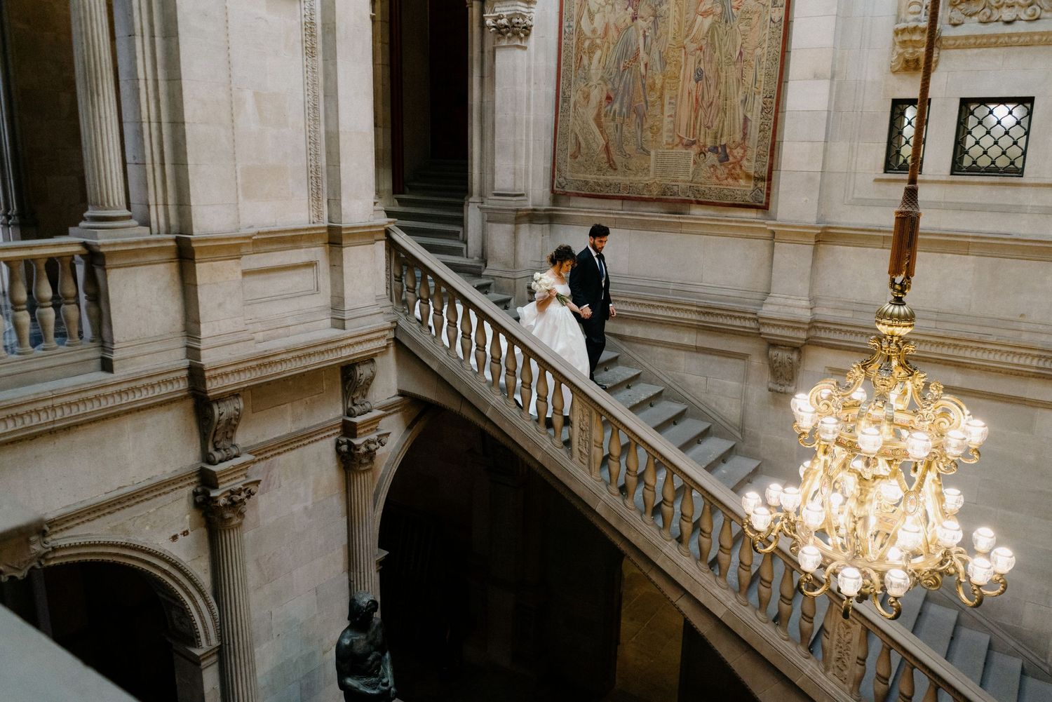 Elegant staircase with chandelier in historic building where couple stands on landing.