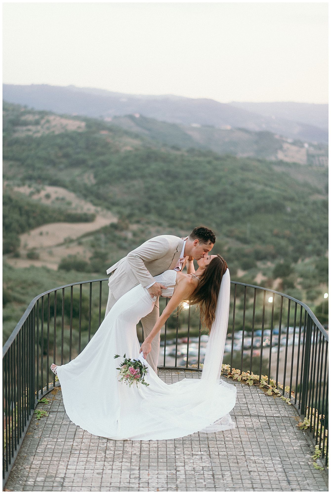Romantic wedding couple sharing a dip kiss on a scenic overlook with rolling hills and mountains in the background.