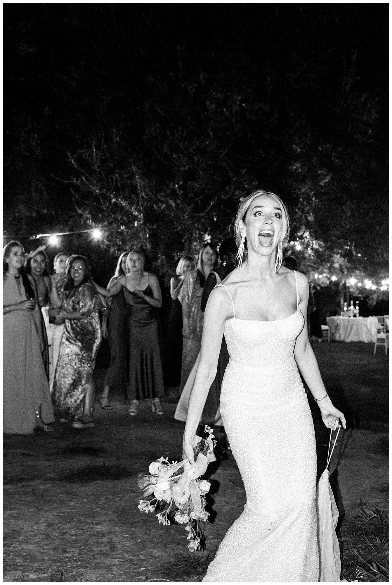 A bride in a fitted white dress holds a bouquet at an outdoor wedding celebration with guests in the background.